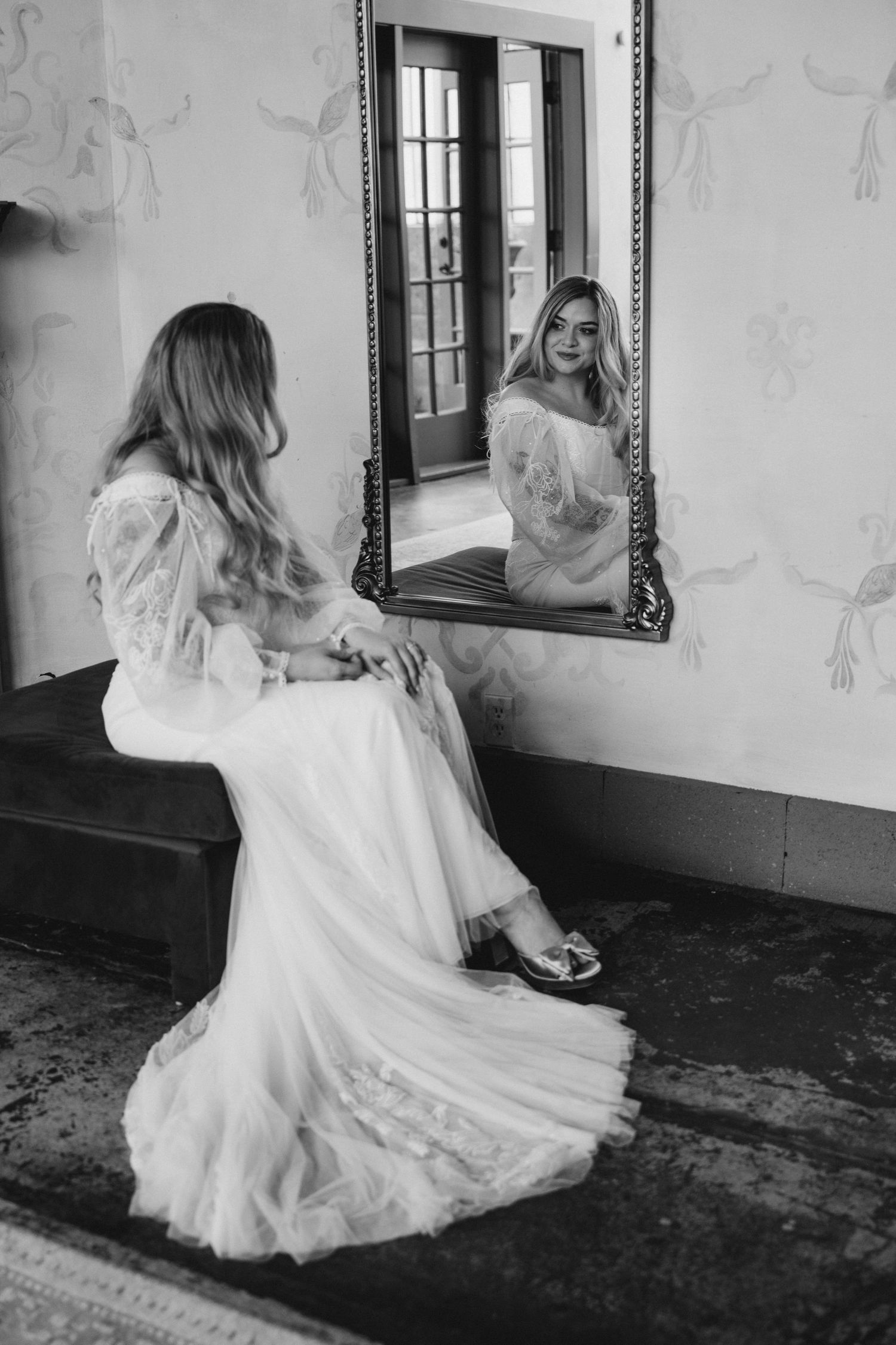 Black and white portrait of bride in lace wedding gown sitting before ornate mirror in elegant room.