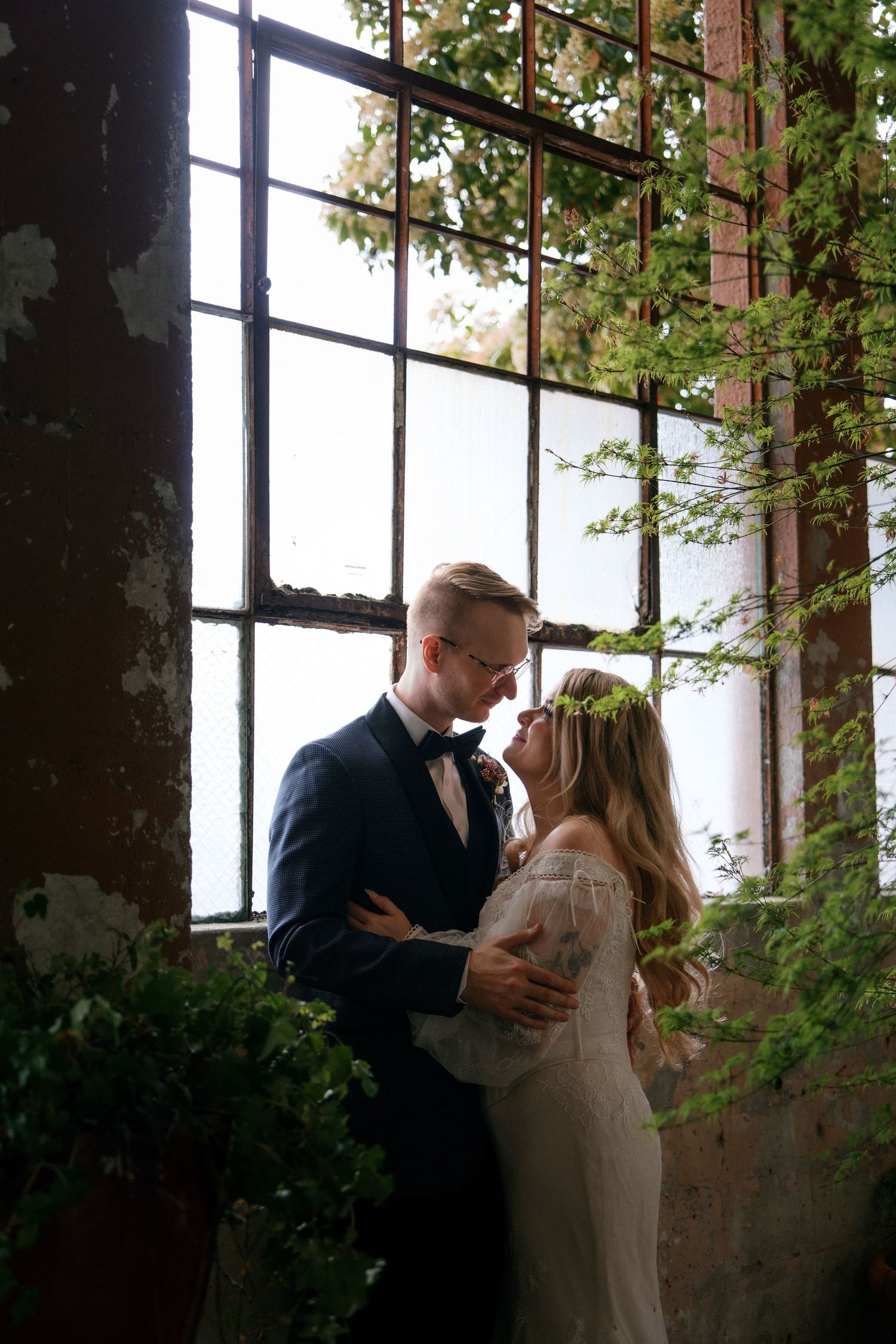 Bride wearing floral crown and groom in navy suit embrace romantically before tall industrial windows with green foliage.