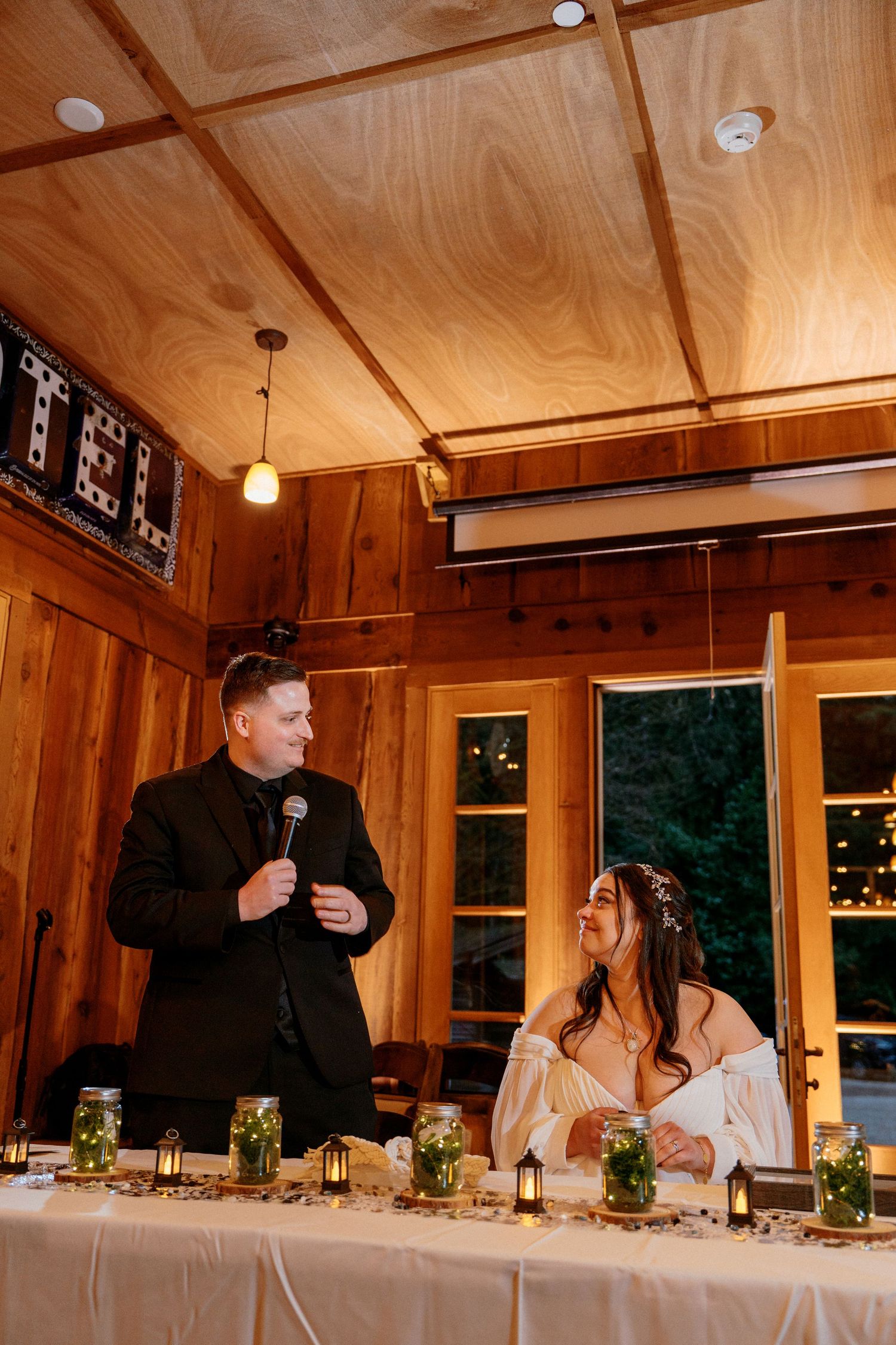 Groom speaks into microphone while bride smiles at rustic wood-paneled wedding reception.