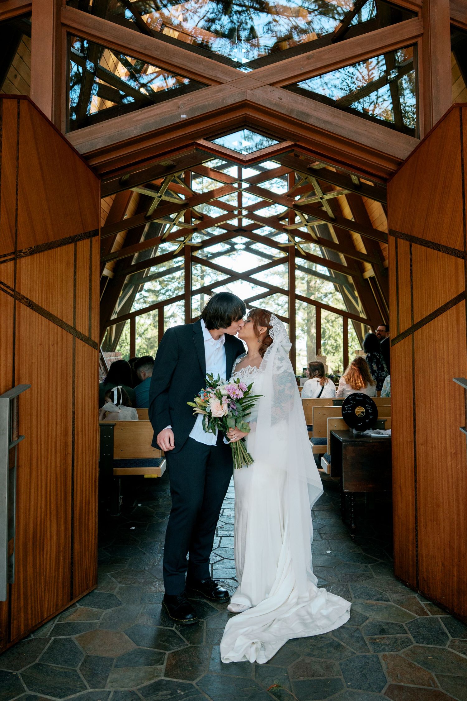 Newlyweds sharing first kiss in a beautiful wood and glass chapel after ceremony.