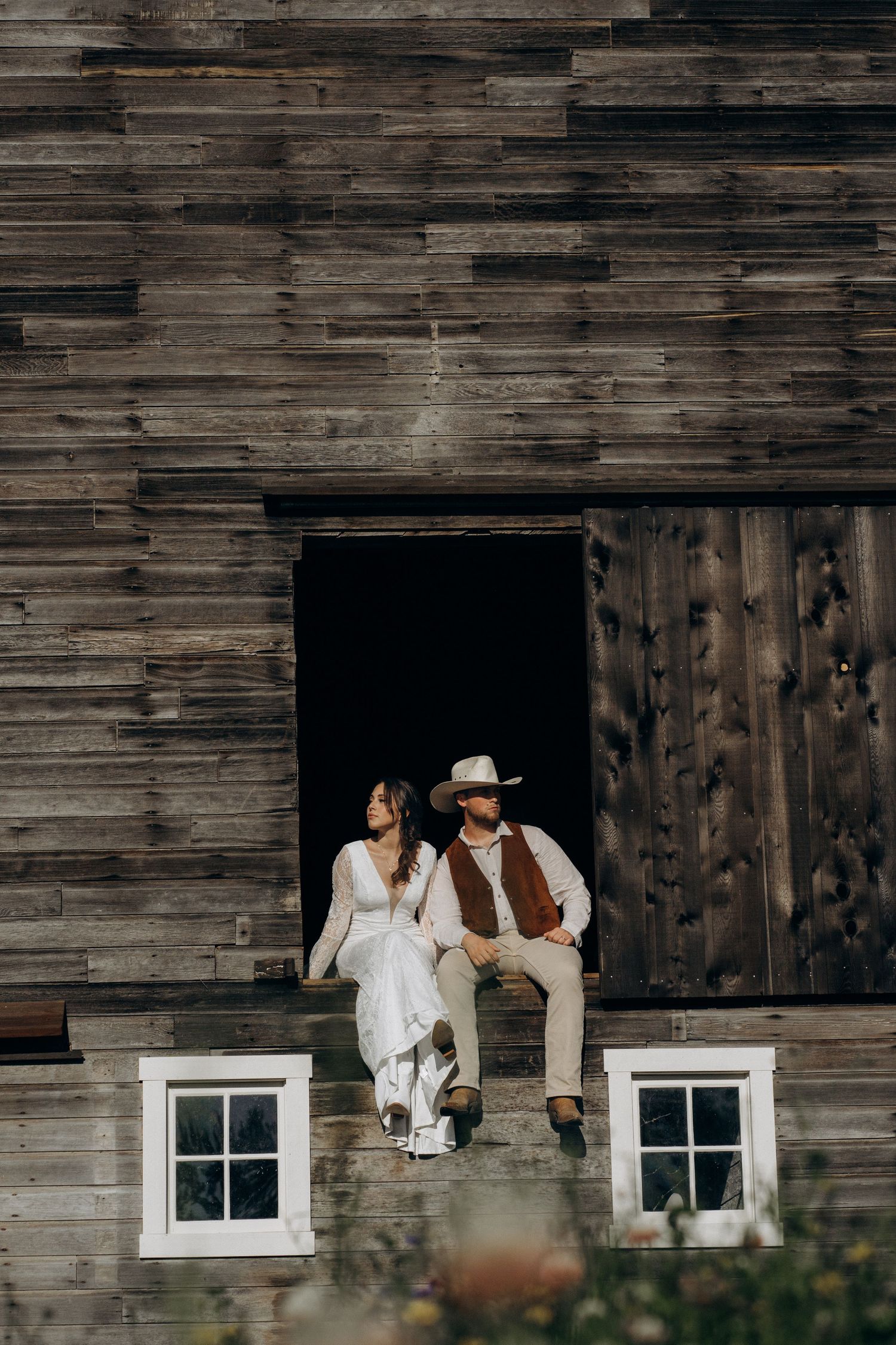 Bride and groom in cowboy hat sit in upper barn doorway above white windows on rustic wooden barn.