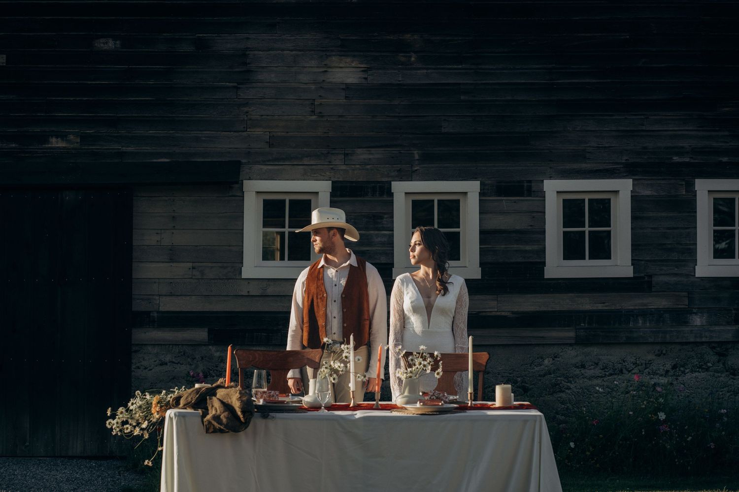 Bride and groom stand at a decorated sweetheart table in front of a rustic dark wood barn at dusk.