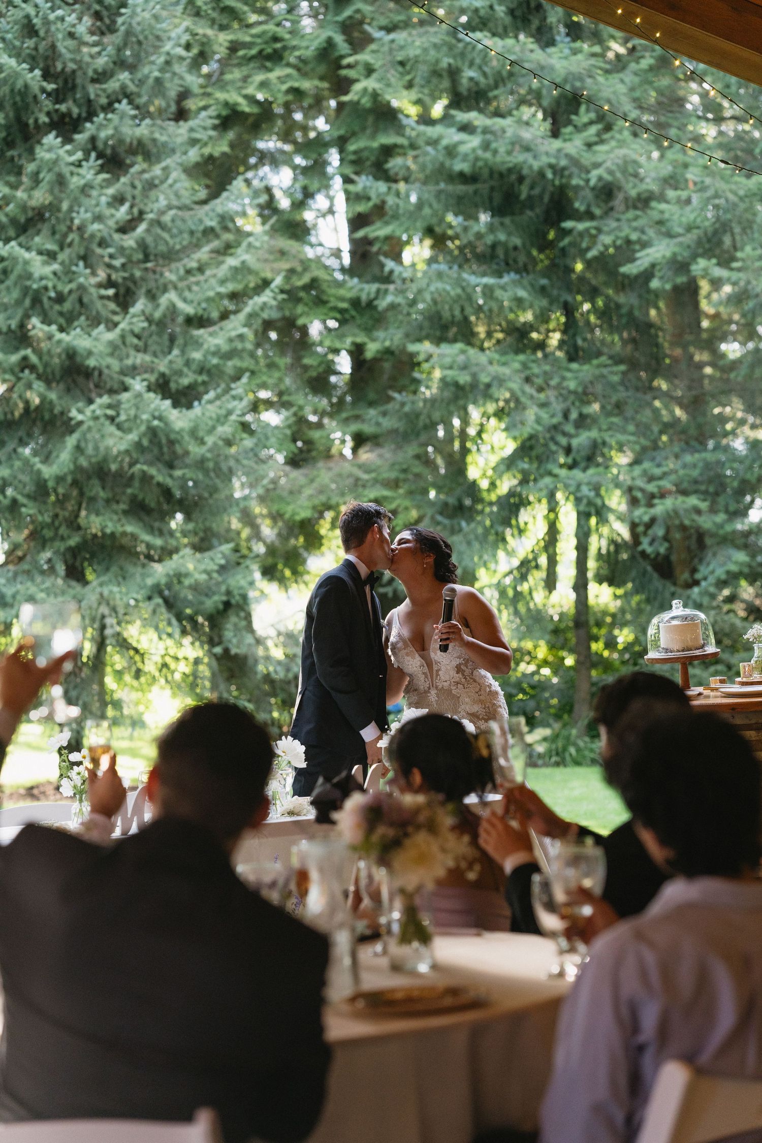 Bride and groom sharing romantic kiss at sweetheart table while wedding guests raise glasses.