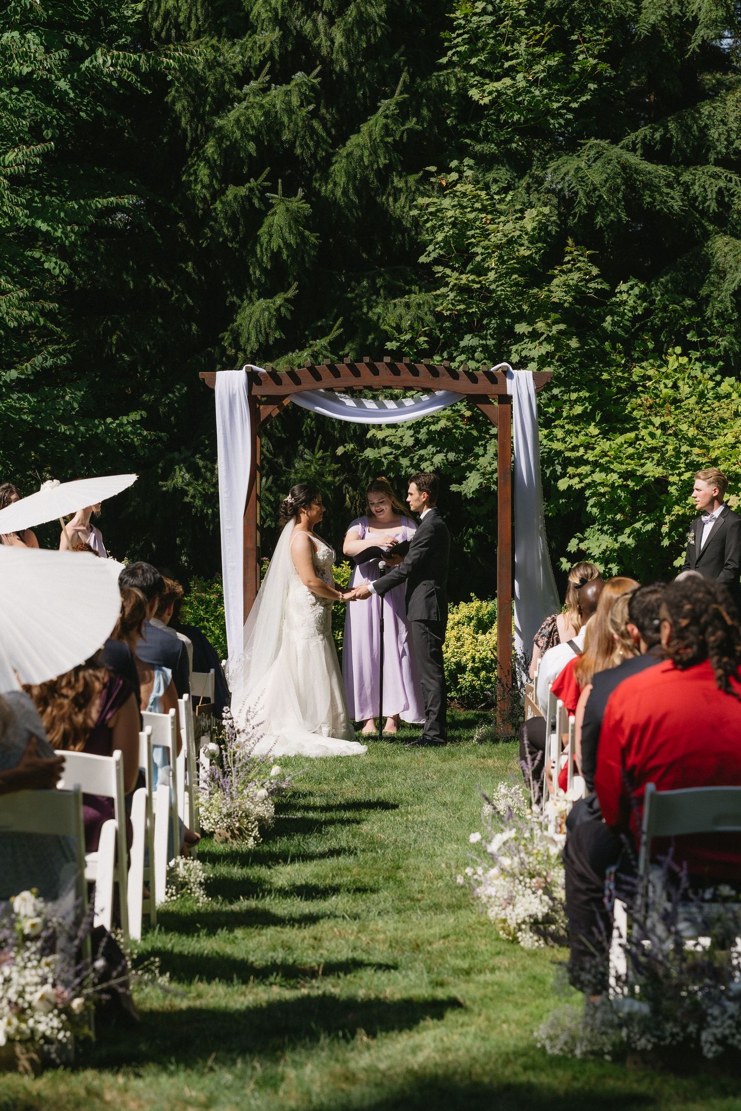 Outdoor wedding ceremony under wooden arch with white draping, bride and groom exchanging vows on green lawn.