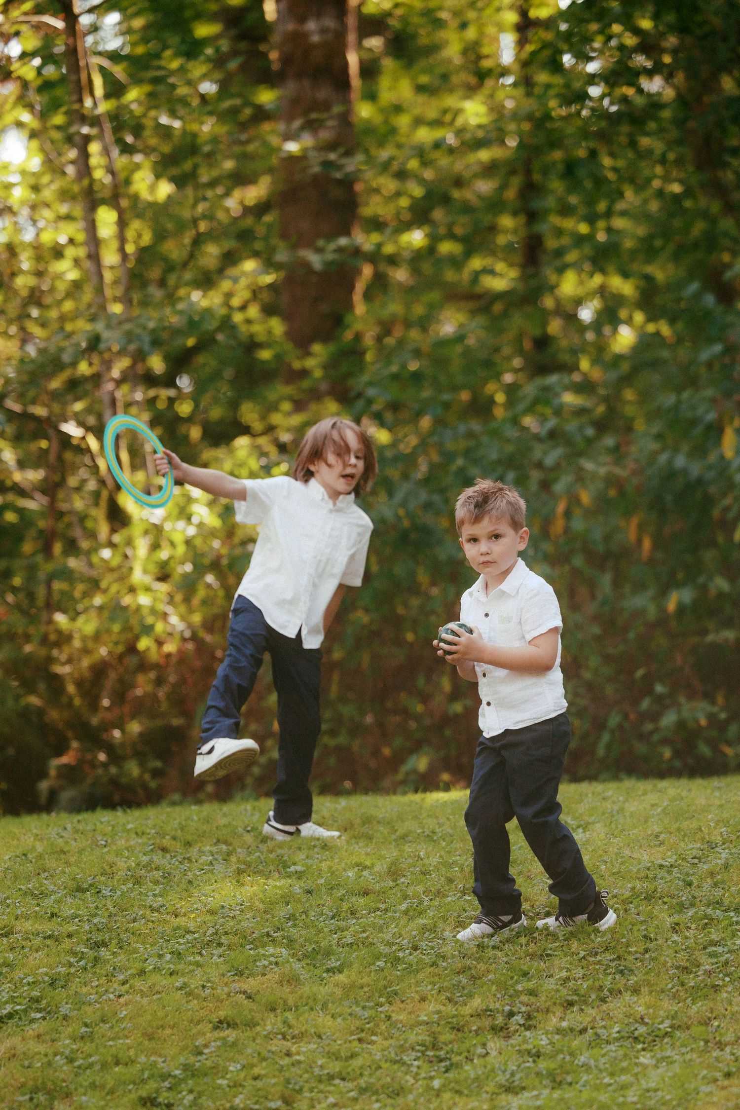 Two young boys in white shirts play with a frisbee ring on green grass at an outdoor wedding.