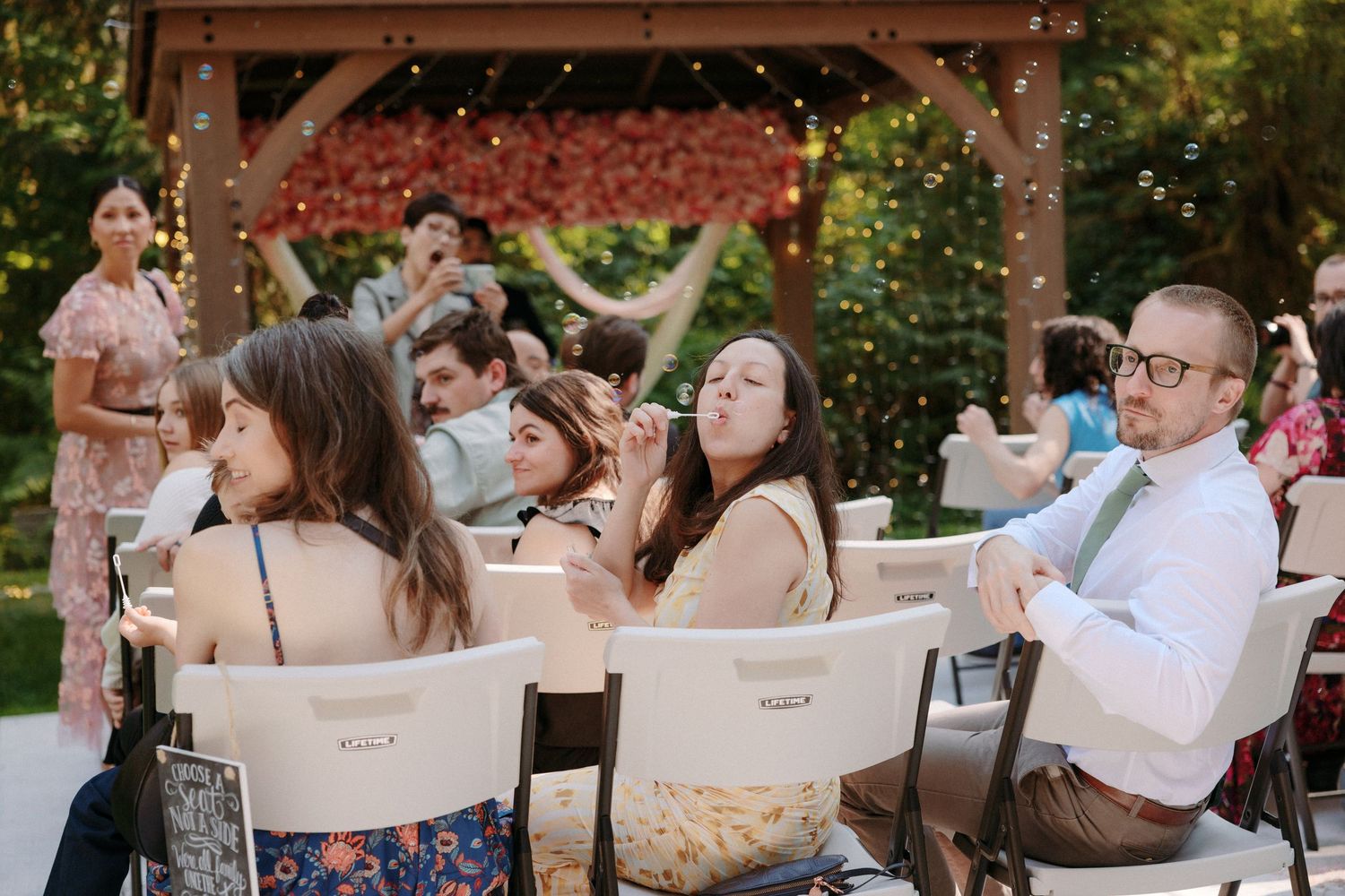 Wedding guests blow bubbles while seated at an outdoor reception under string lights.