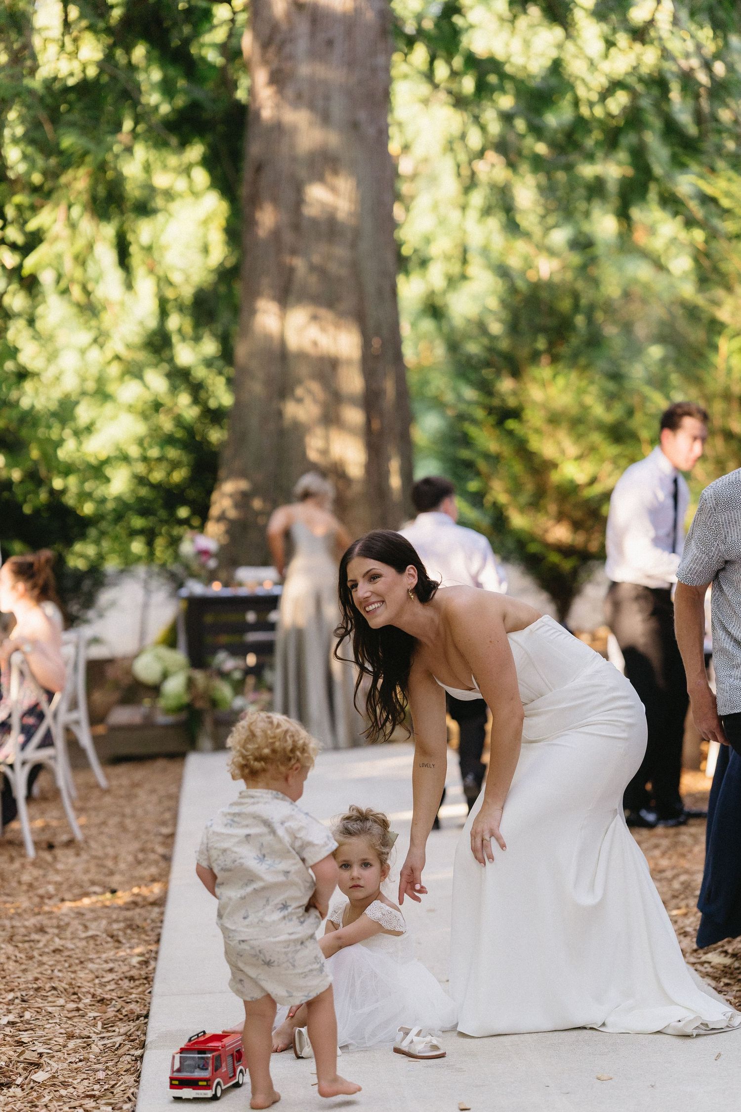 Bride in white gown bending down to interact with two young children on a garden path.