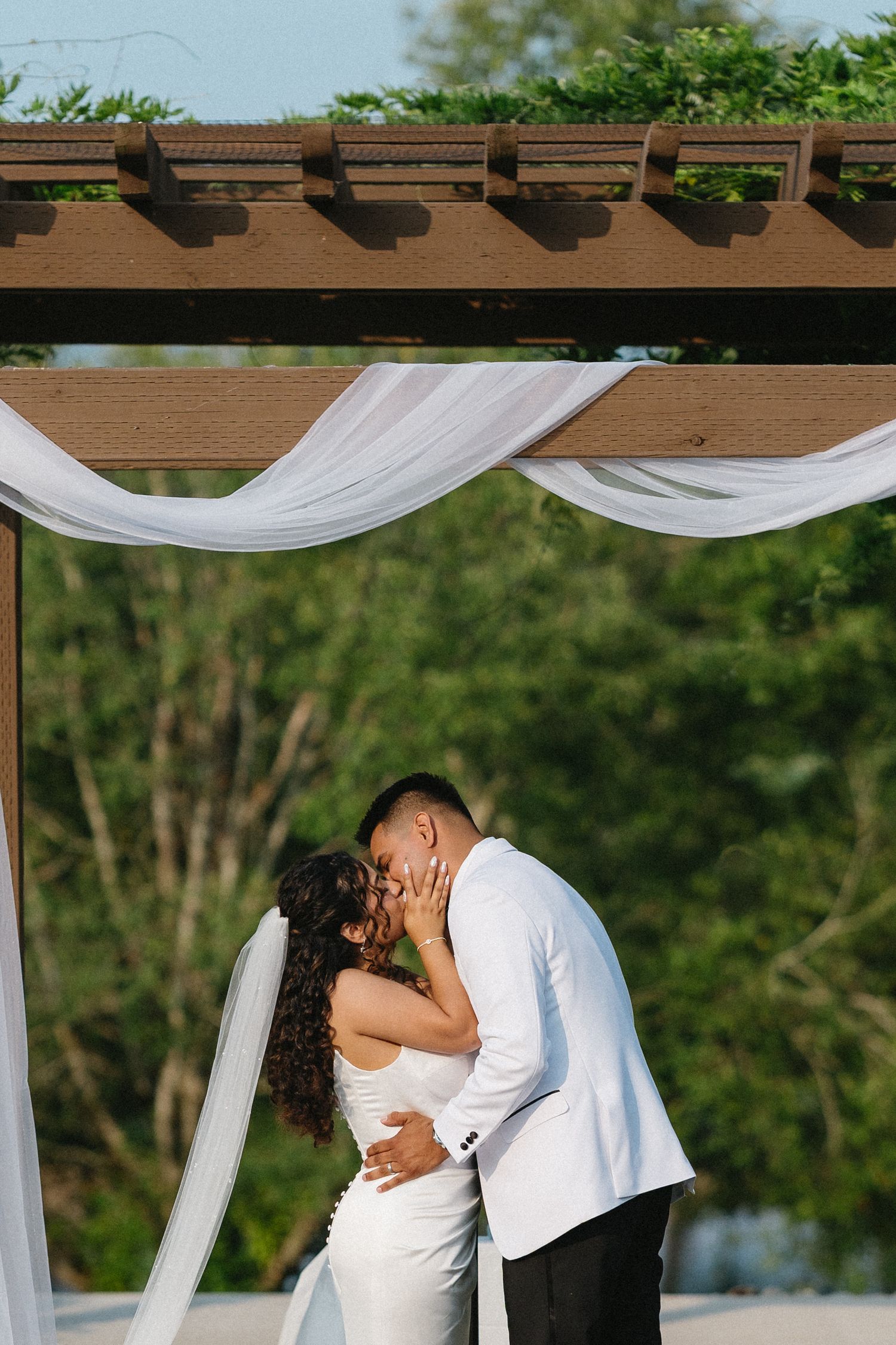 Bride and groom share first kiss under draped pergola at outdoor wedding.