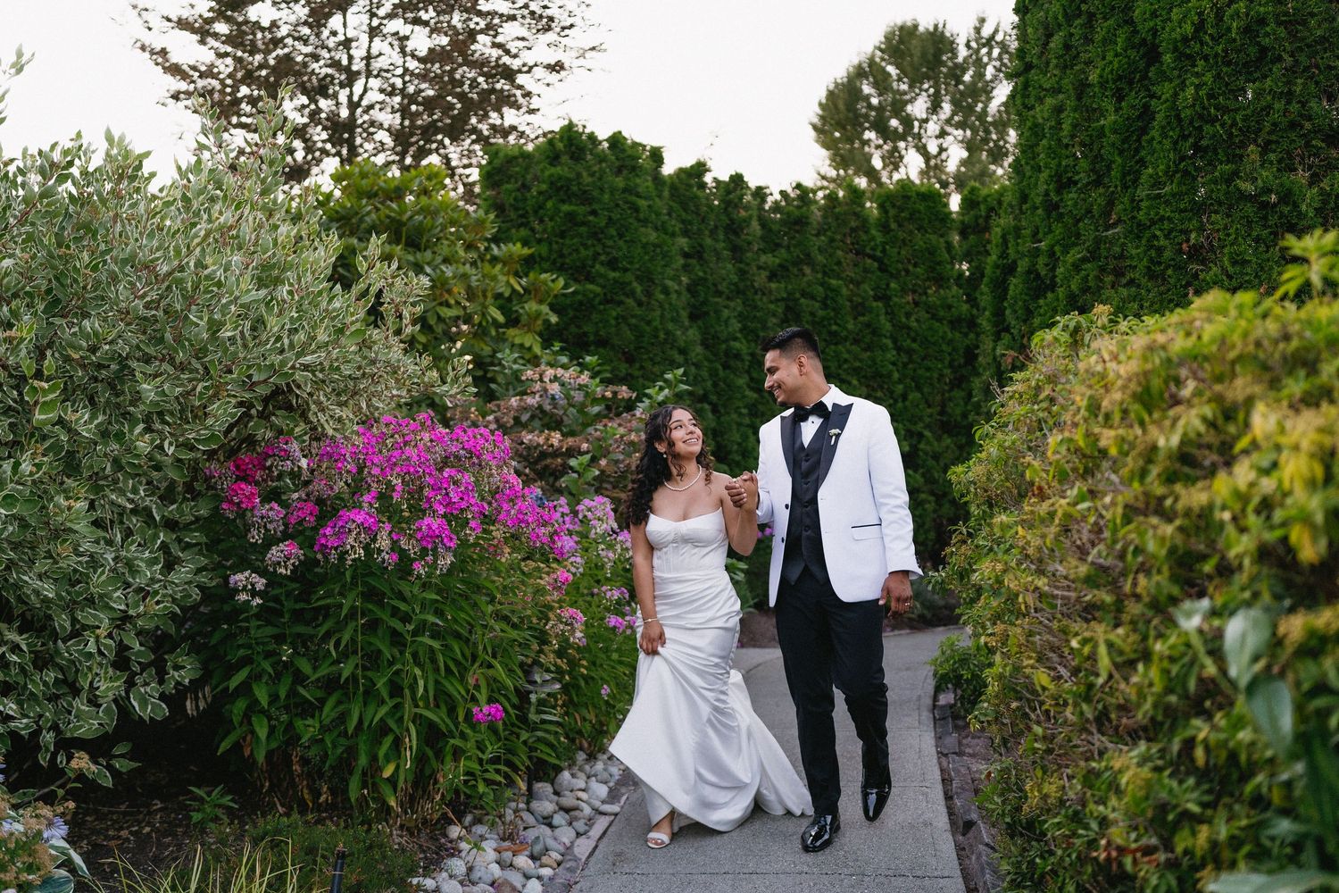 Bride and groom walking hand in hand on a garden path surrounded by lush greenery and pink flowers.