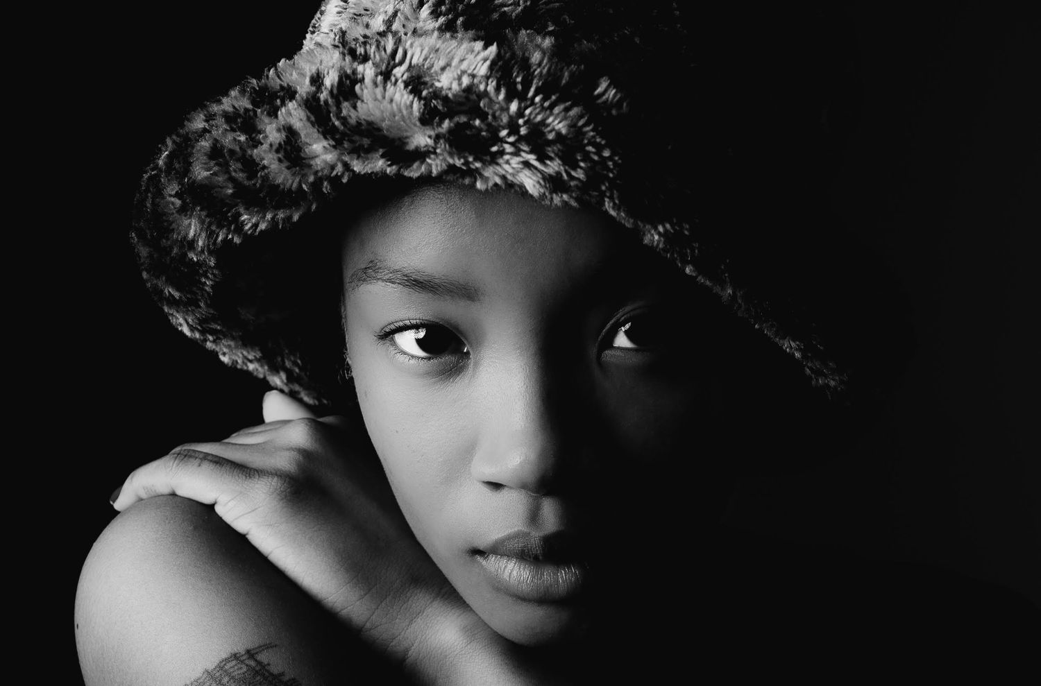 Black and white close-up portrait of a young person wearing a stylish fur hat against a dark background.