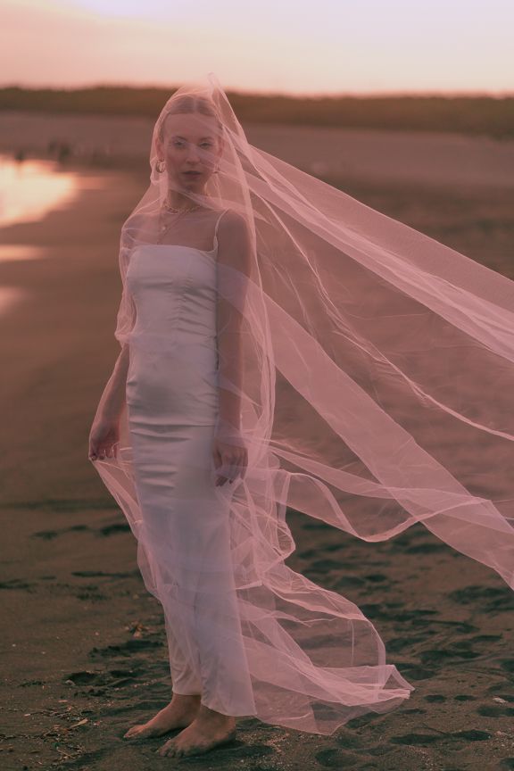 Bride in white gown with flowing veil stands on sandy beach at golden sunset.