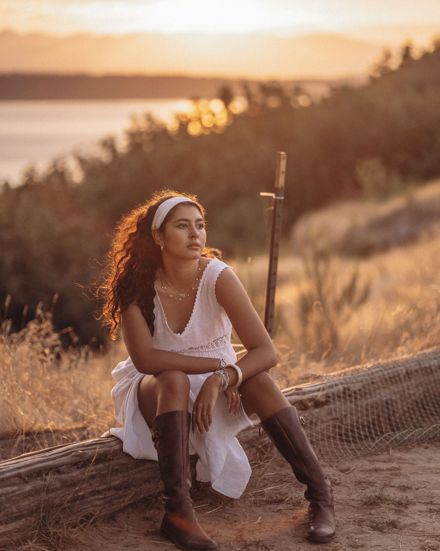 Woman in white dress and boots sits on log at golden sunset with sword nearby.