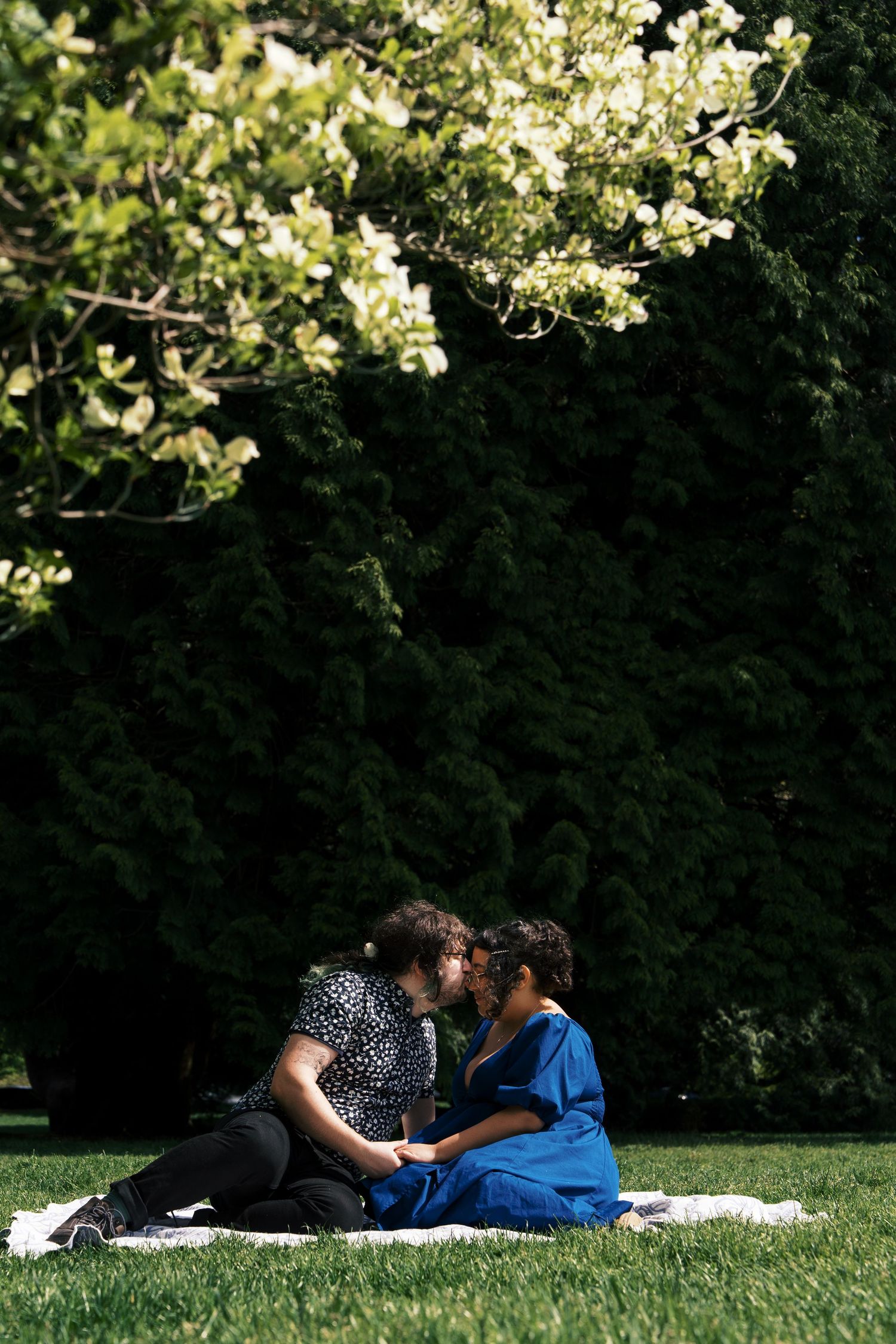 Couple sharing an intimate moment on a white blanket beneath flowering trees in a sunlit garden.