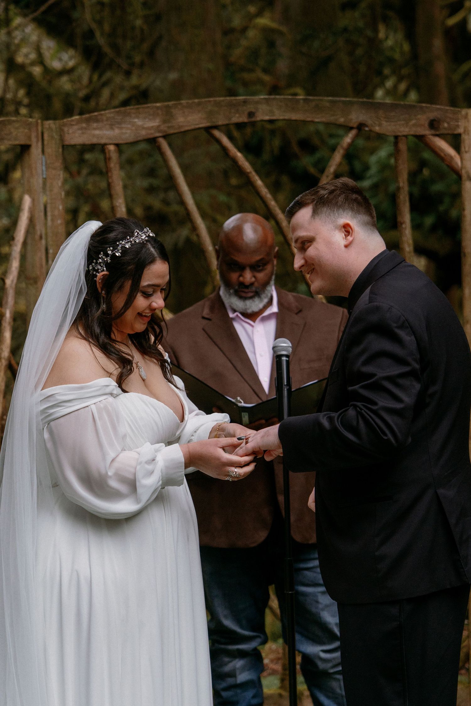 Bride and groom exchanging rings outdoors under rustic wooden arch with officiant during wedding ceremony.