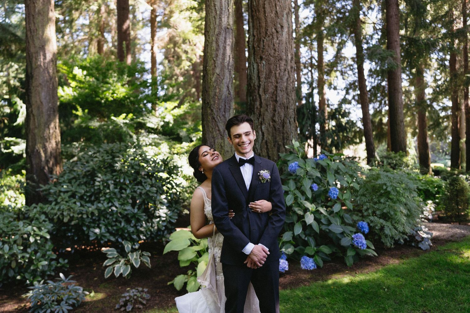 Bride and groom laughing together among tall trees and blue hydrangeas in an outdoor garden.