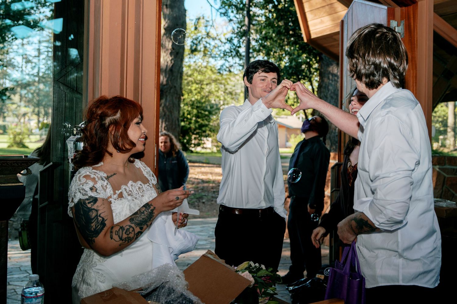 Tattooed bride in lace wedding dress laughing with guests at an outdoor wedding reception.