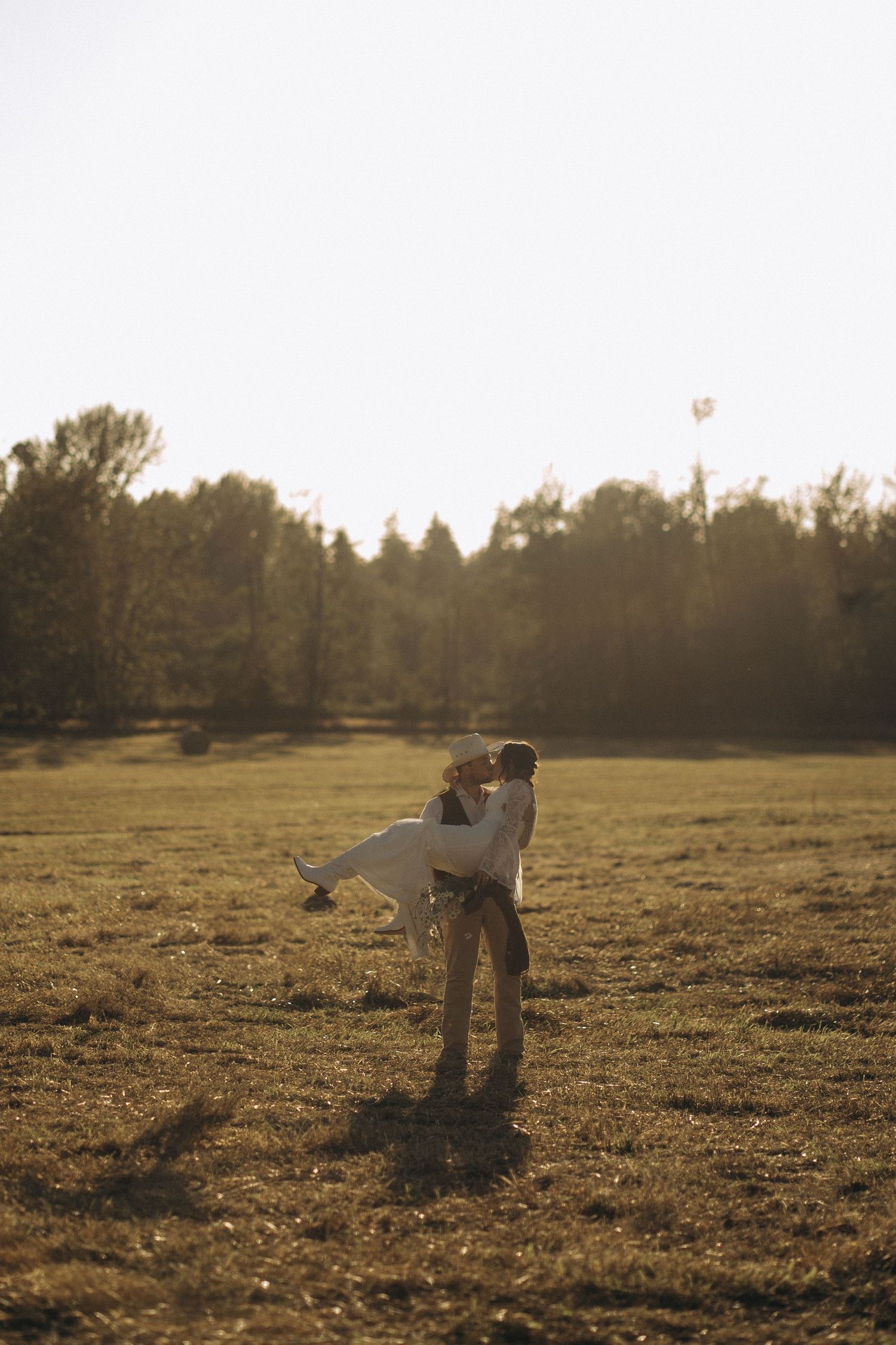 Groom lifting bride in sunlit open field during romantic outdoor wedding photo session.