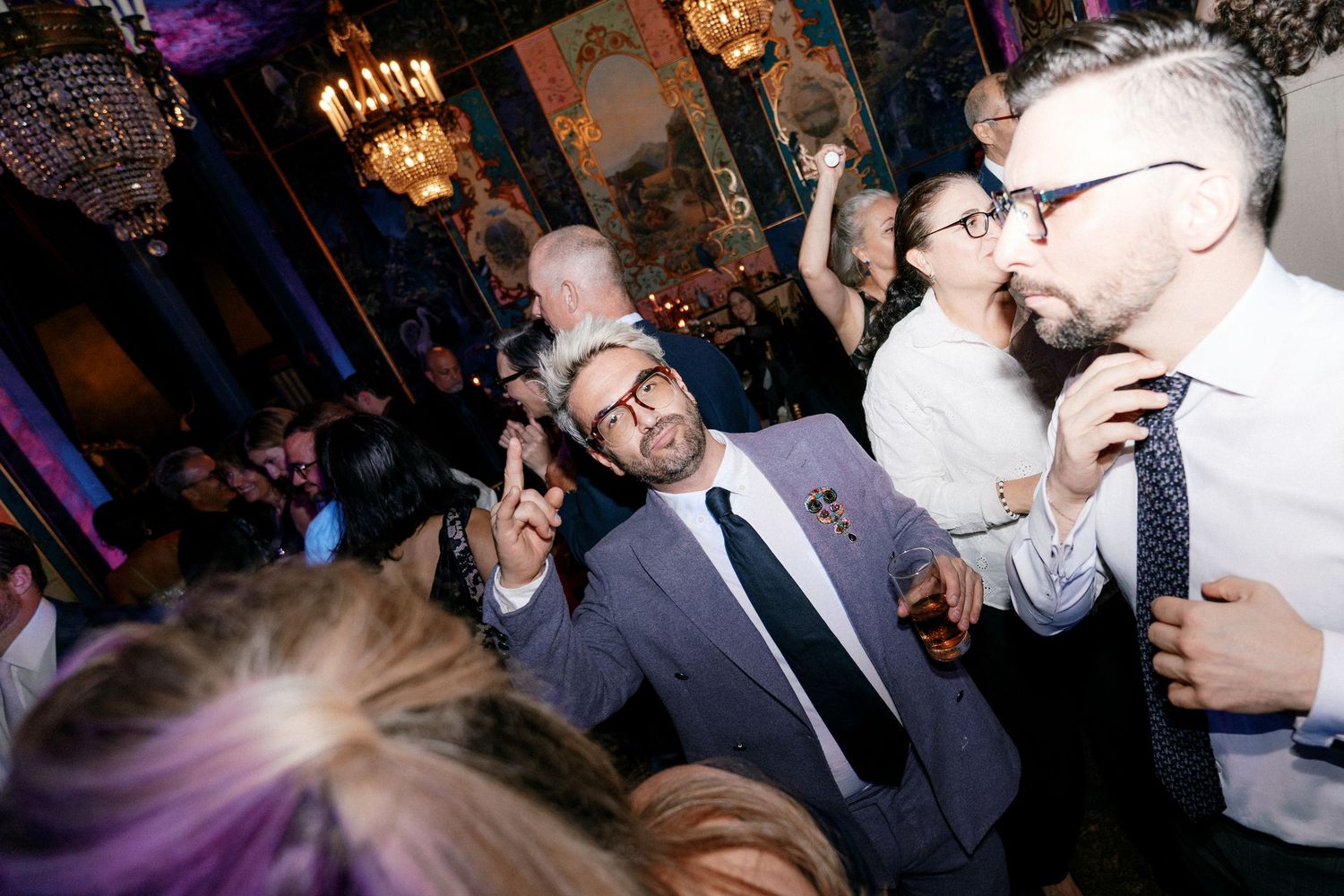Man in gray suit smiling and gesturing at lively wedding reception party.