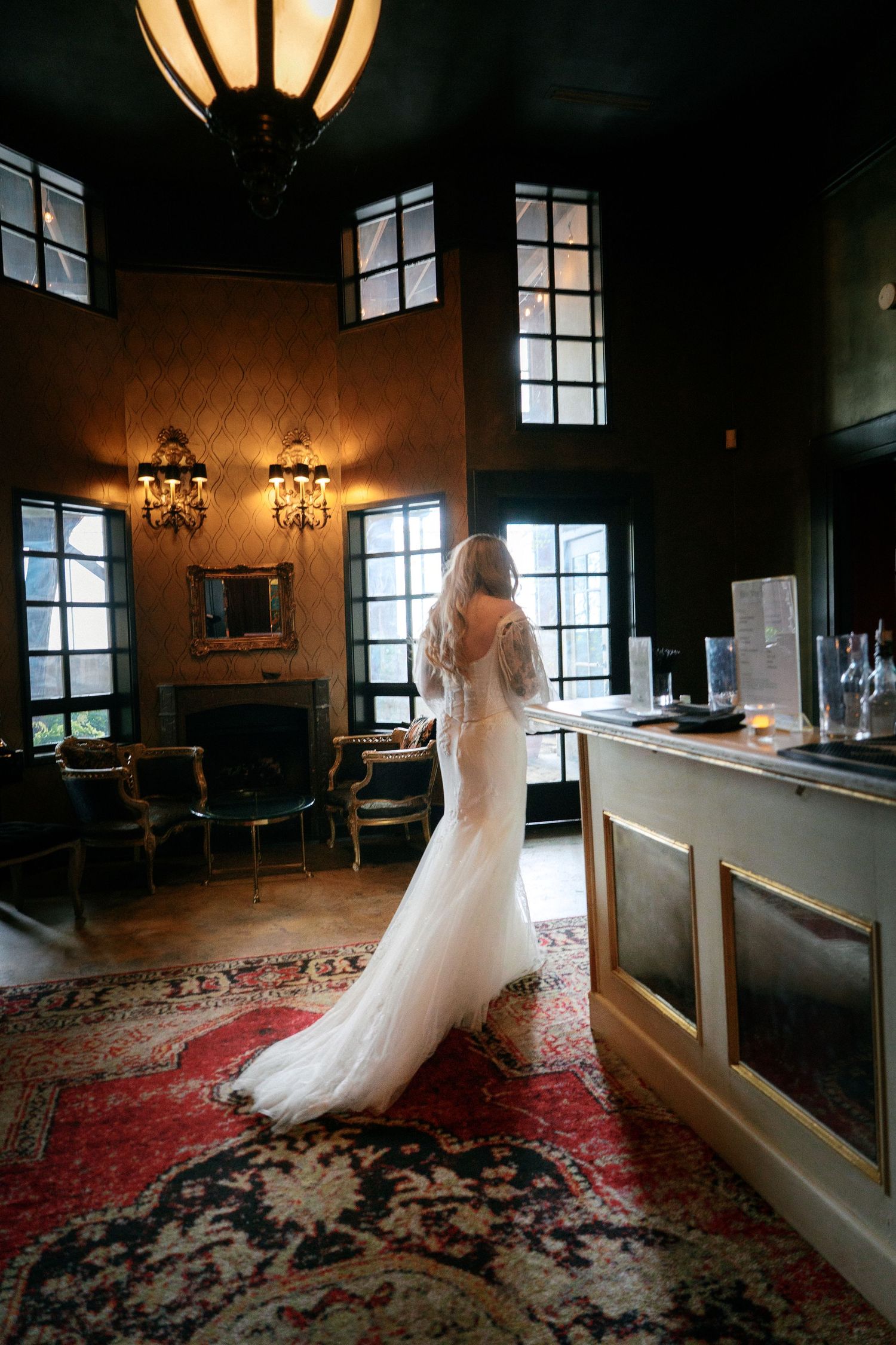 Bride in a fitted white gown with long train standing at a bar in a dimly lit elegant venue.