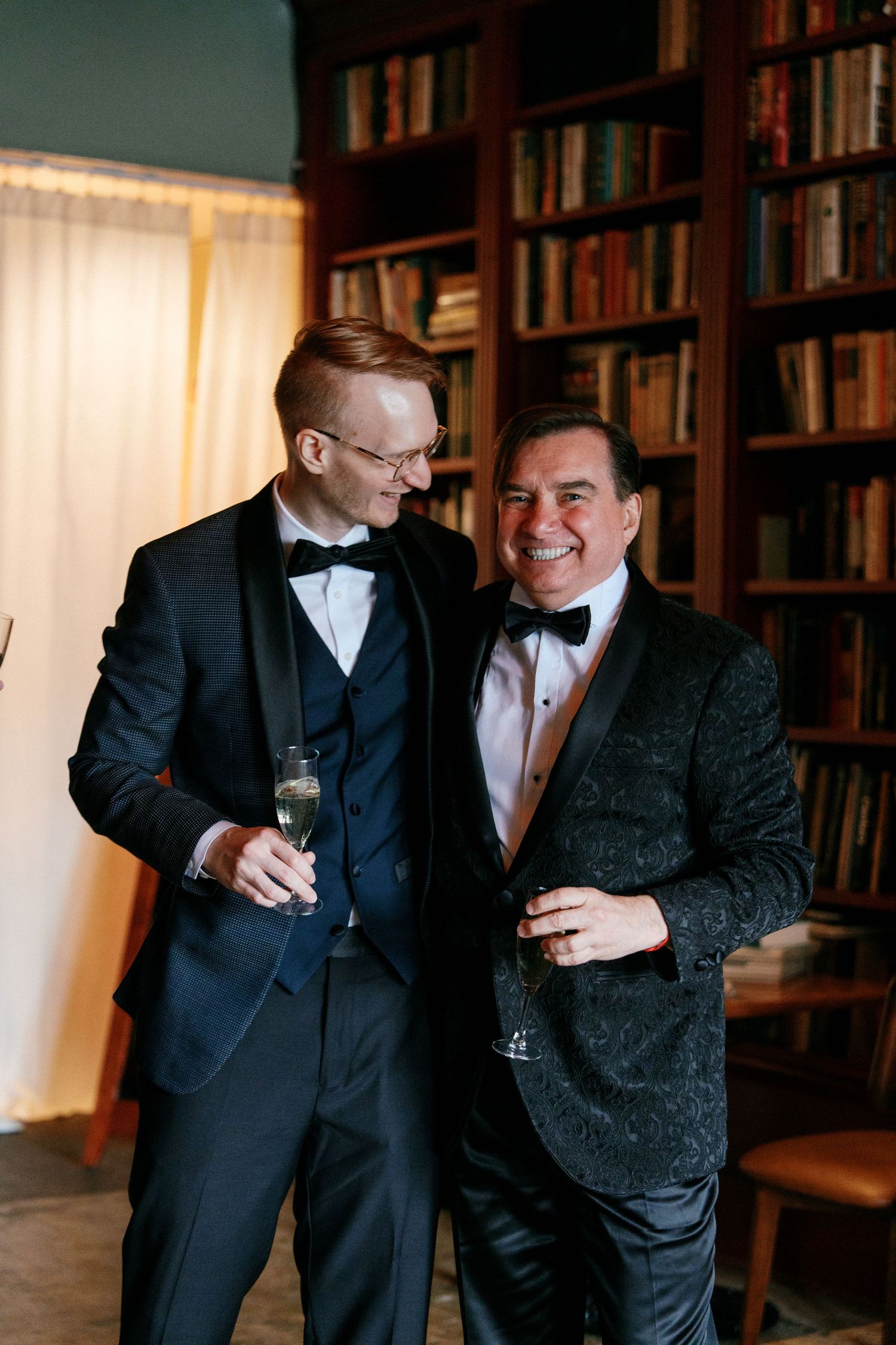 Two men in black tuxedos smiling and holding champagne glasses in front of bookshelves.