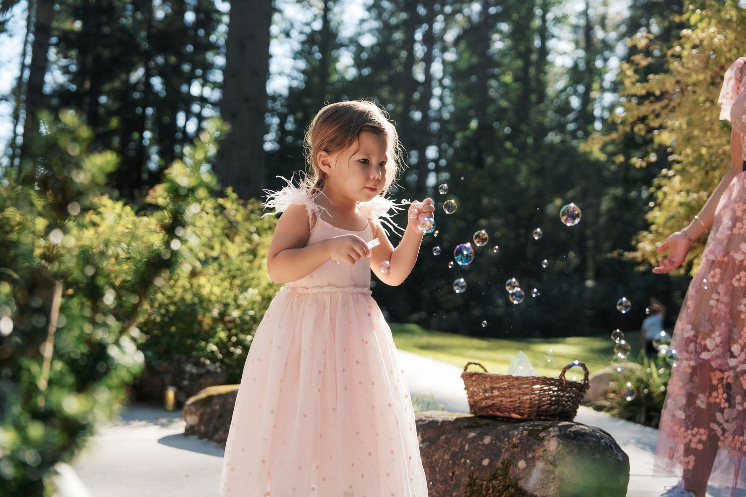 Young girl in pink tulle dress blowing bubbles outdoors at a sunny garden wedding celebration.