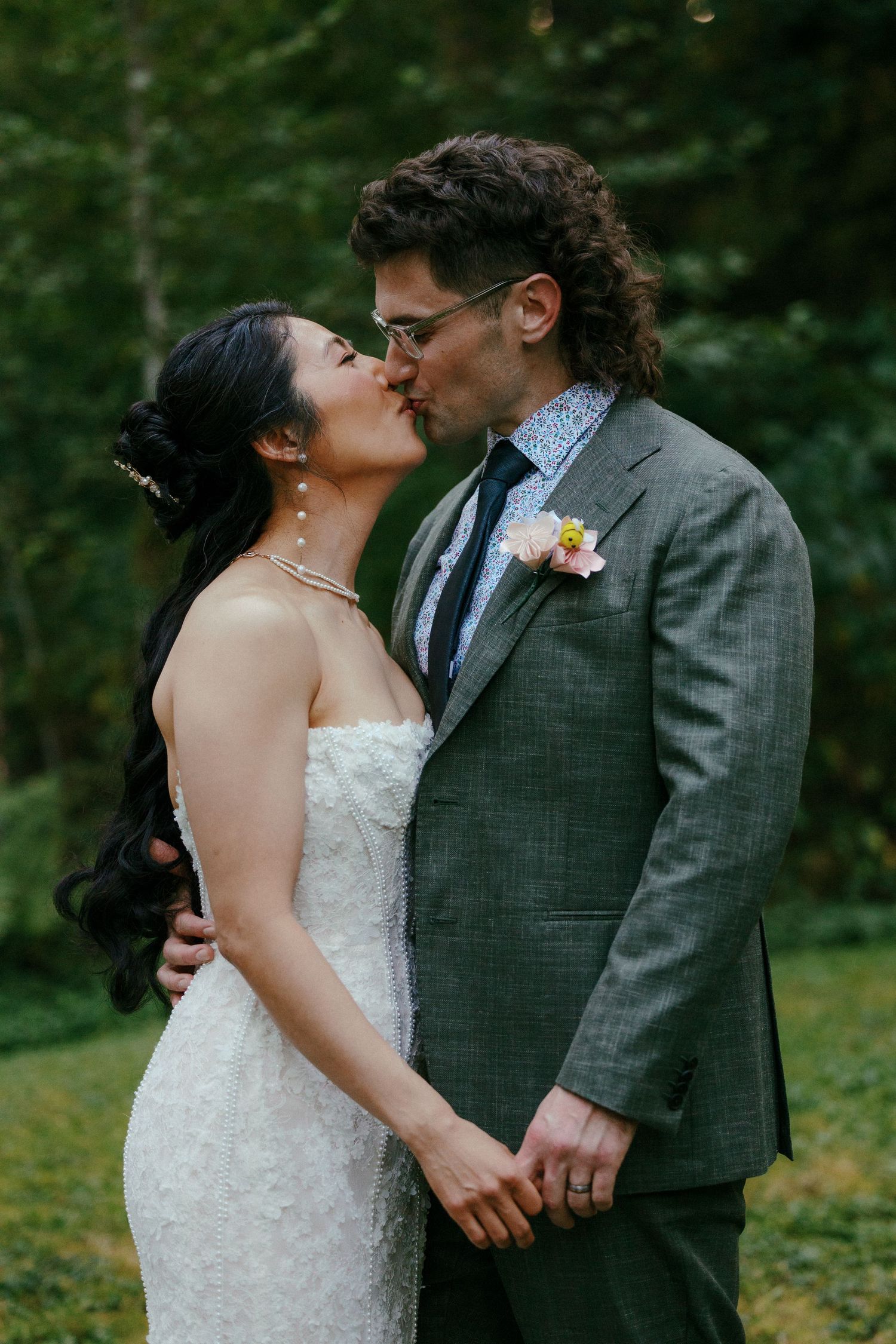 Bride and groom share a romantic kiss outdoors, groom wearing gray suit with floral boutonniere.