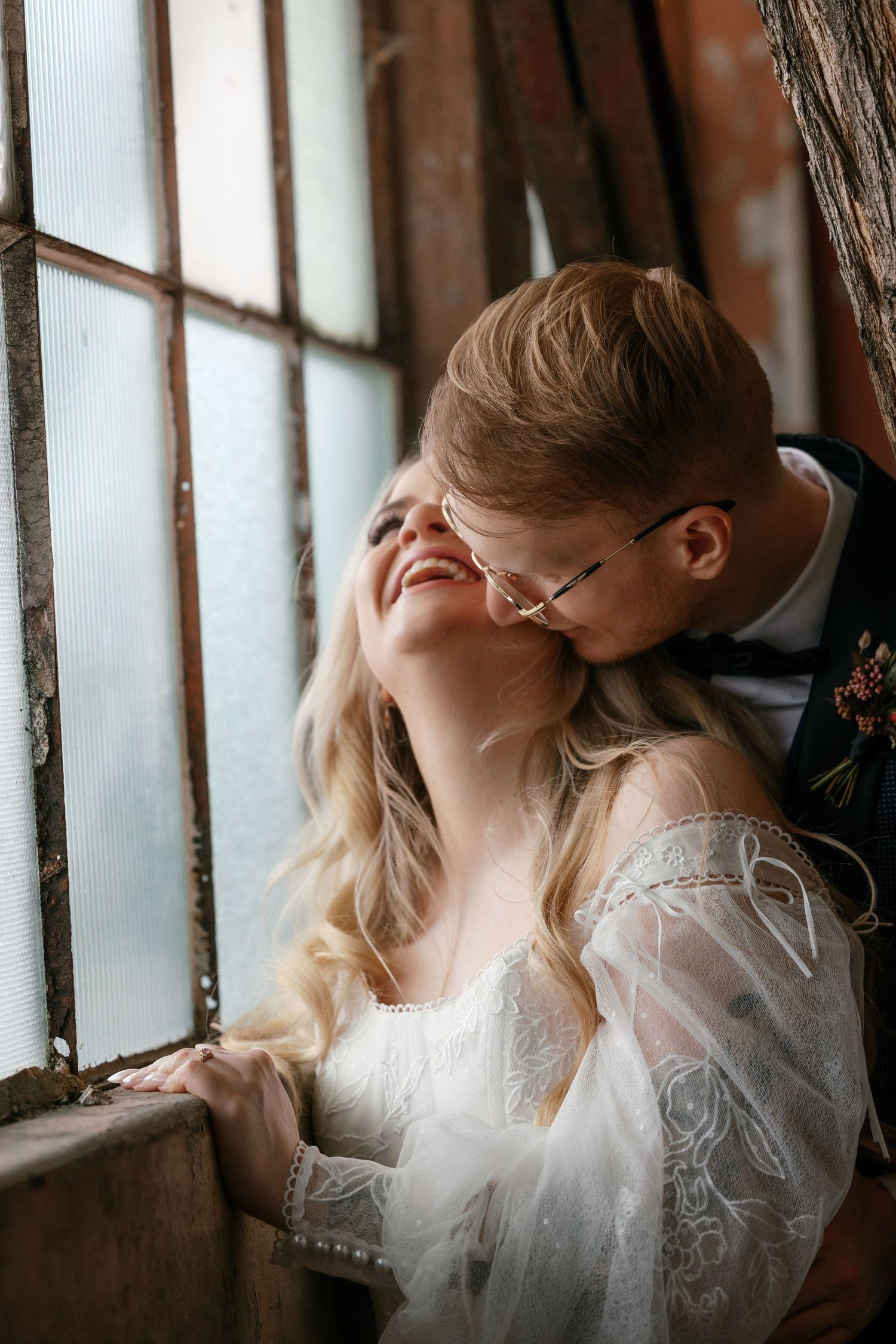 A groom kissing his smiling blonde bride on the cheek near a rustic window, both dressed in elegant wedding attire.