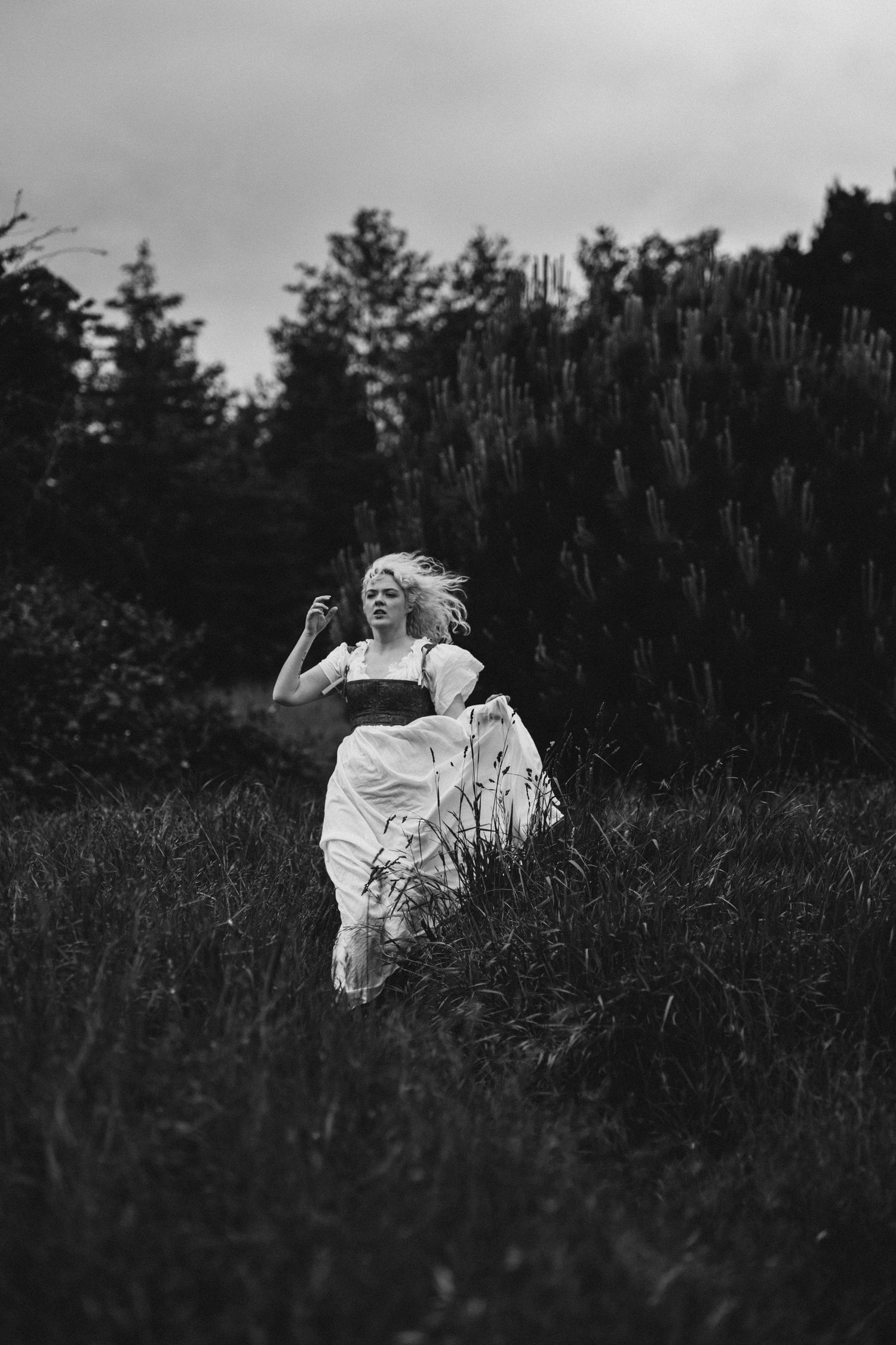 Black and white photo of a blonde woman in a white dress running through a dark, moody field with trees in the background.