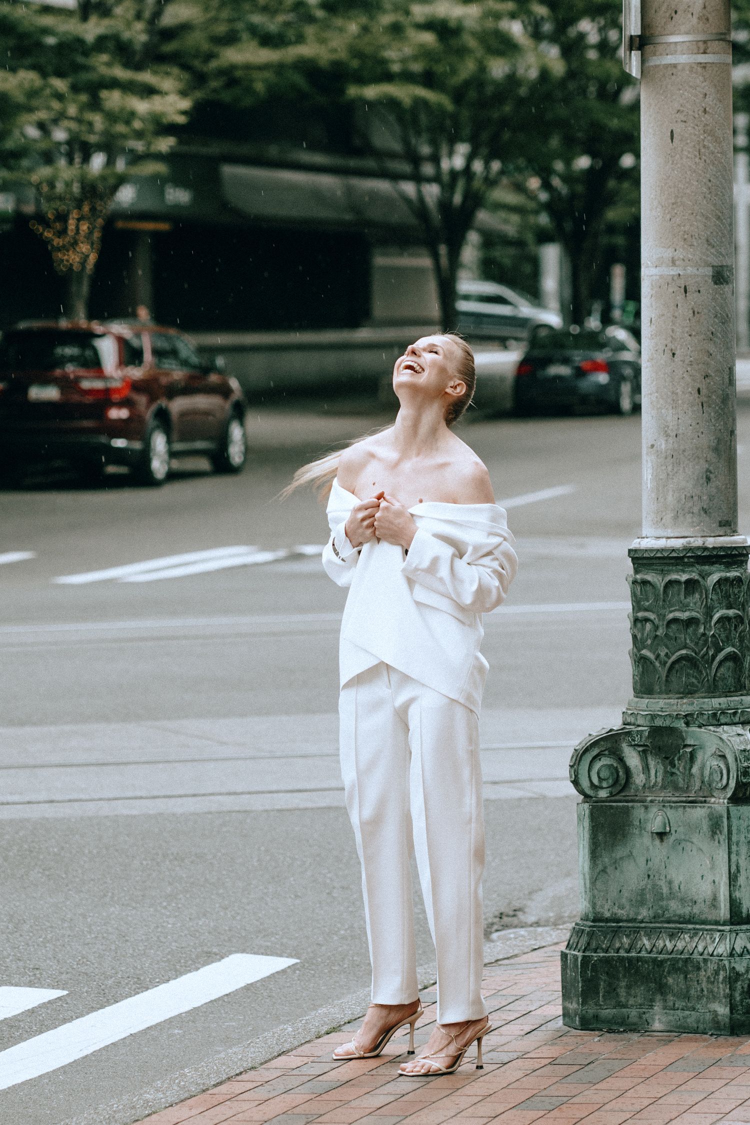 Blonde woman in white off-shoulder blazer laughs joyfully beside decorative street lamp at urban intersection.