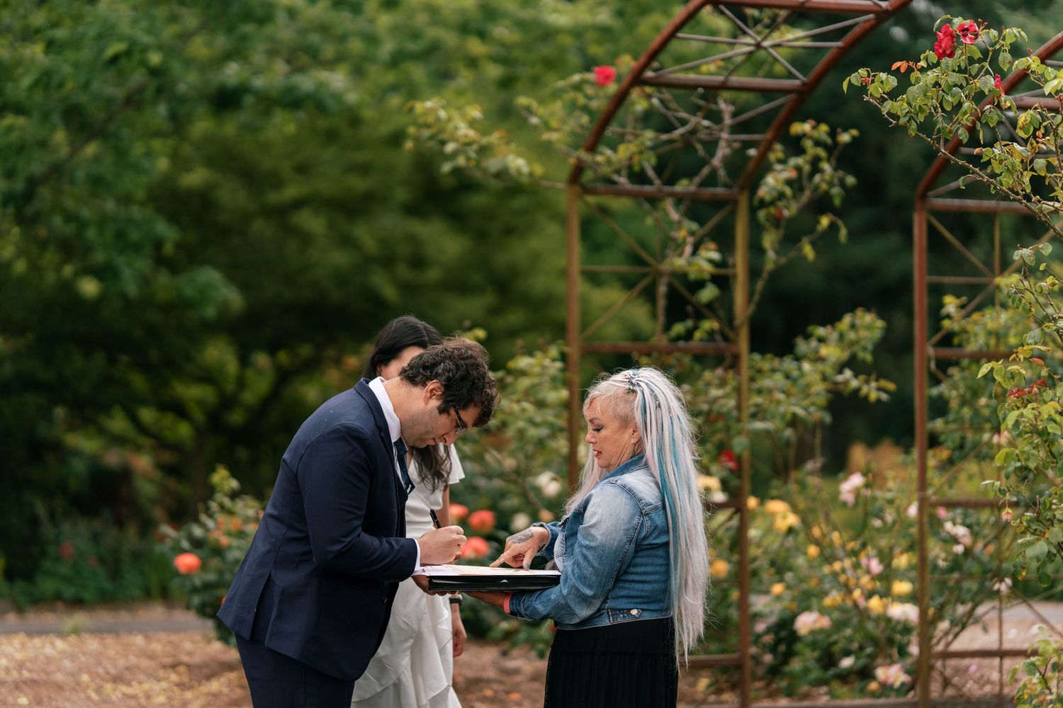 Groom signing marriage certificate with officiant in a lush rose garden setting.