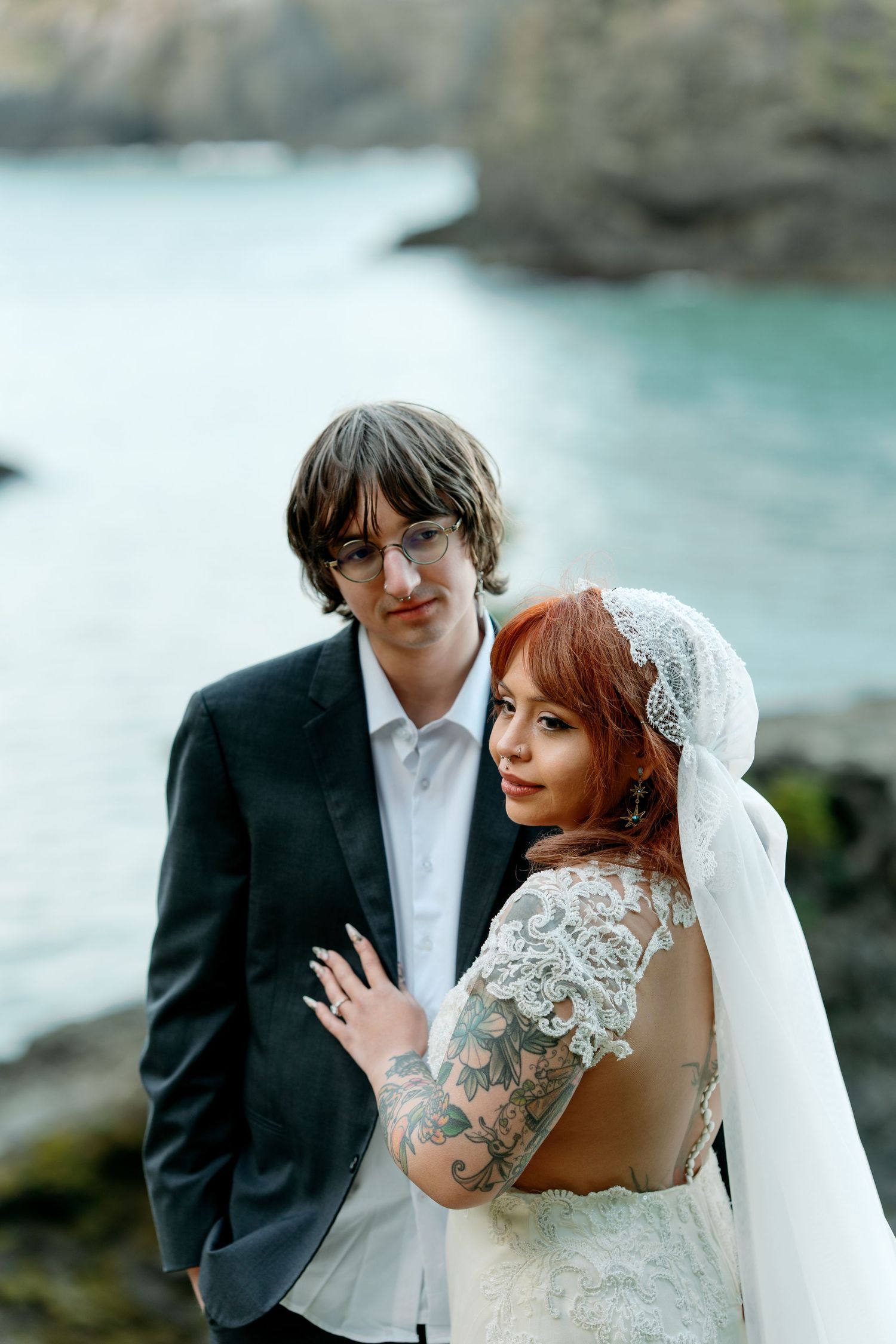 Tattooed redhead bride in lace gown and groom in dark suit pose together by rocky coastal waters.