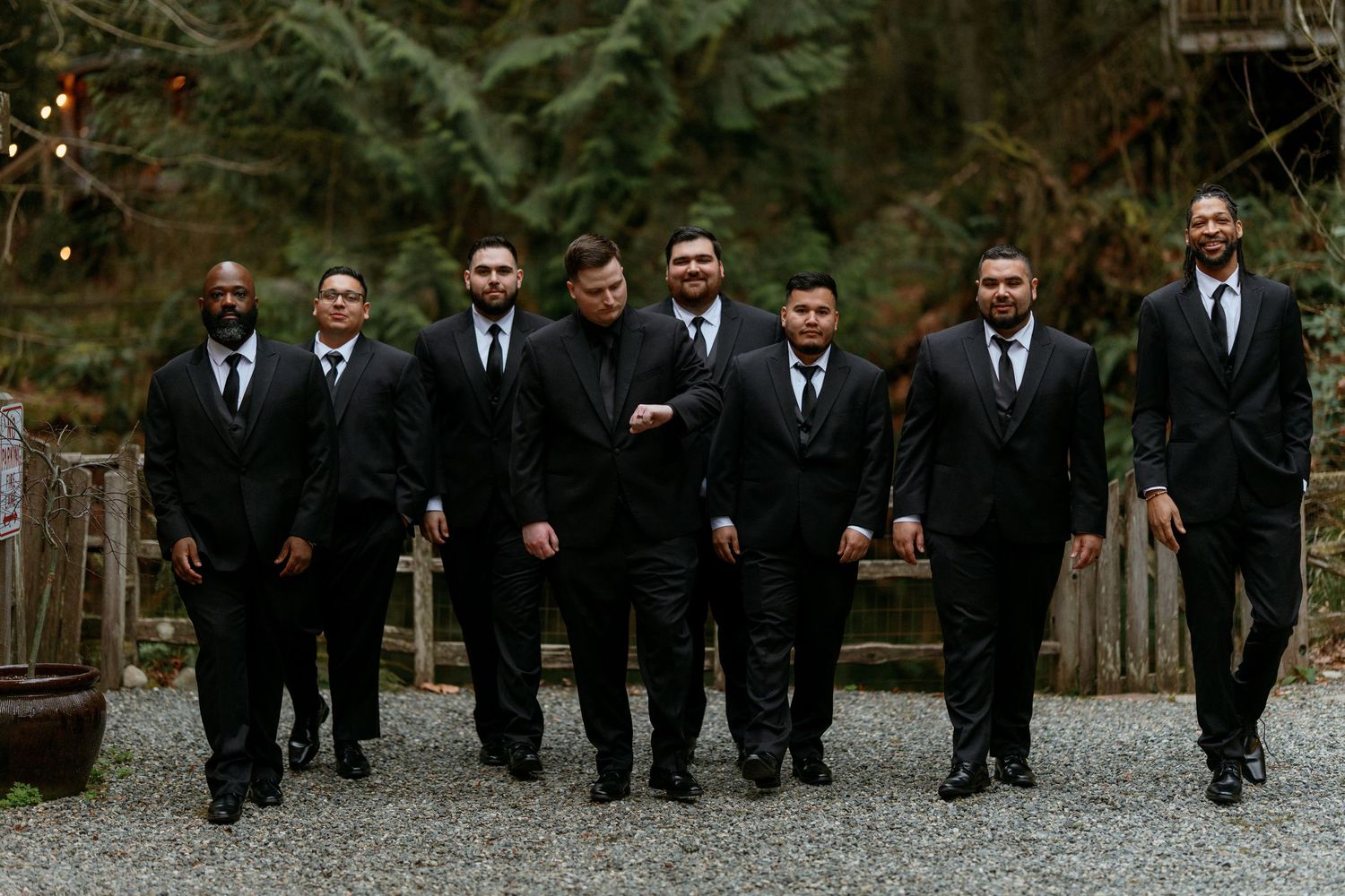 Eight groomsmen in black suits walking confidently at outdoor forest wedding venue.