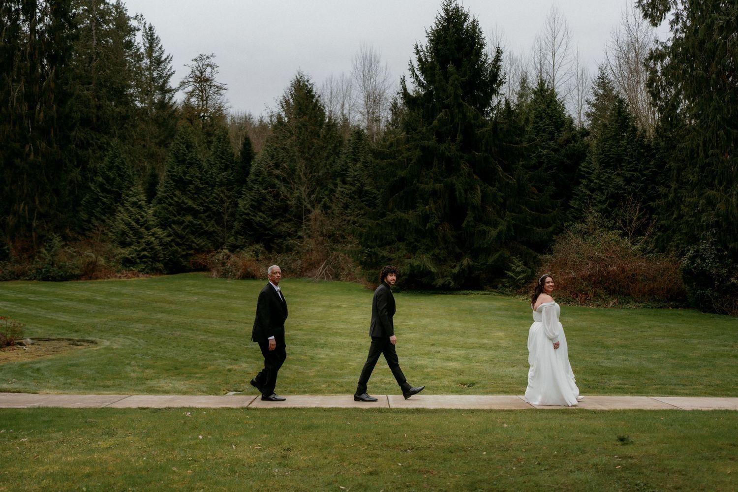 Three wedding guests walk along garden path surrounded by evergreen trees at outdoor wedding venue.