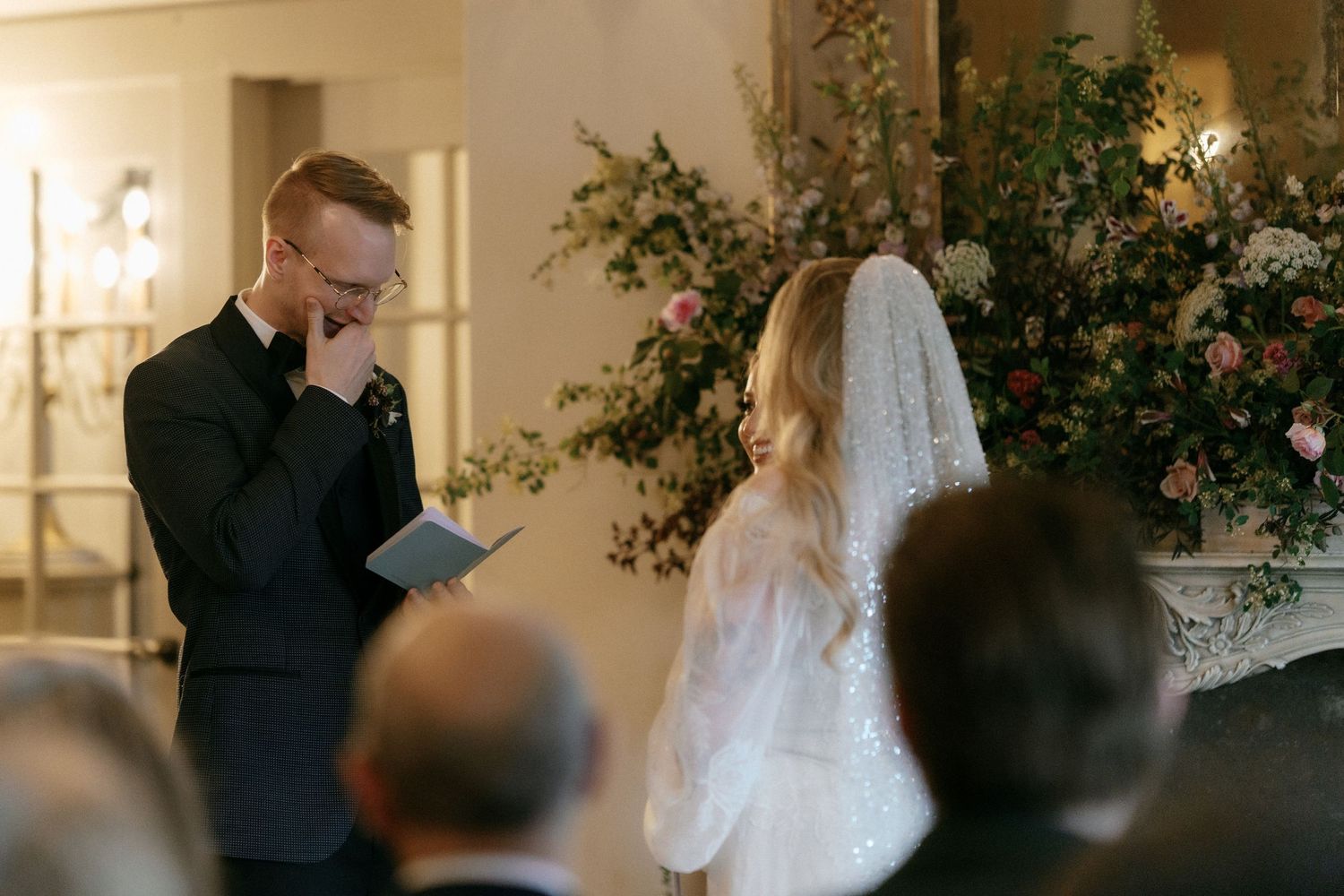 Groom reads emotional wedding vows while bride stands before floral backdrop at elegant indoor ceremony.