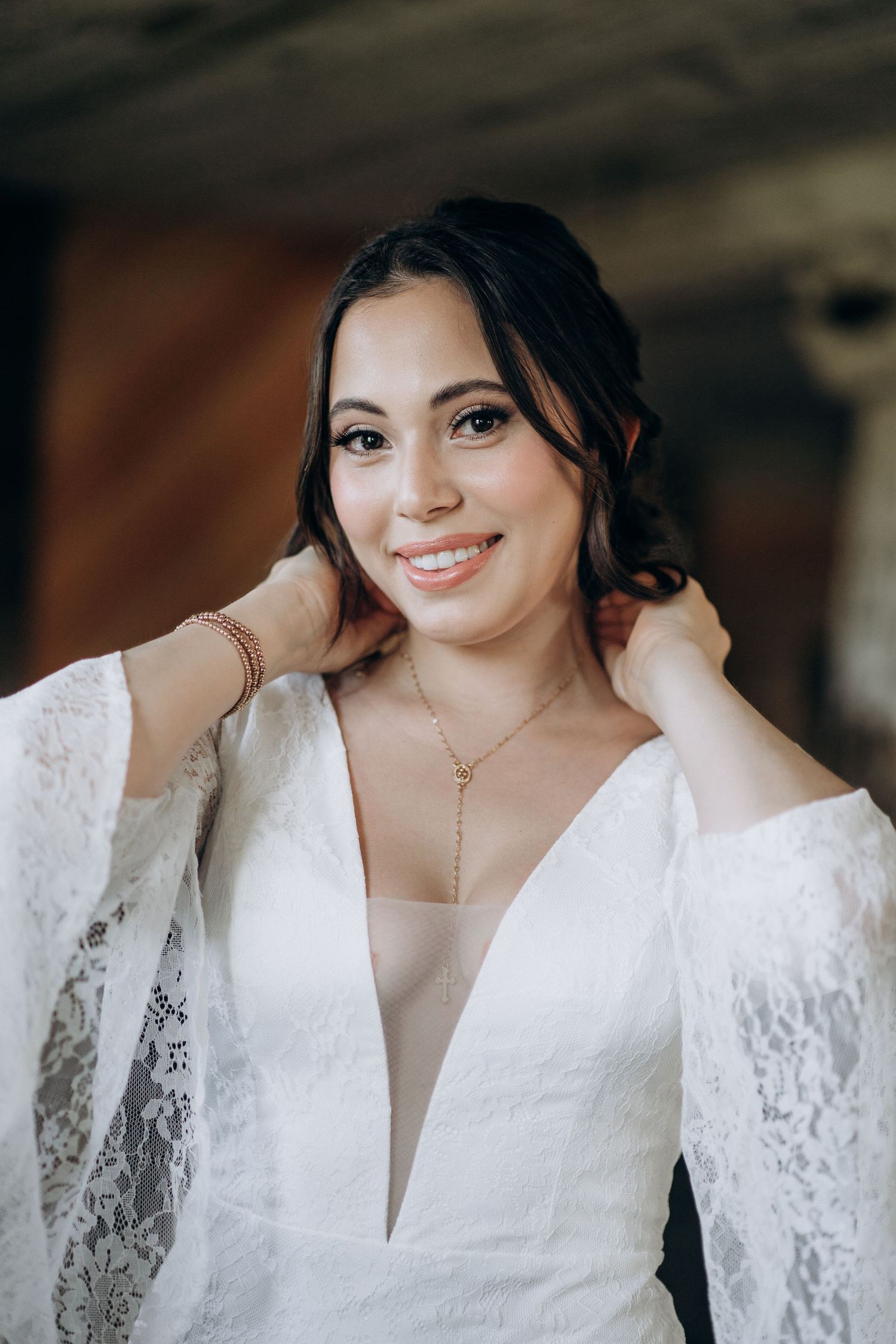 Radiant bride in deep V-neck white lace dress adjusting hair, wearing gold necklace in rustic setting.