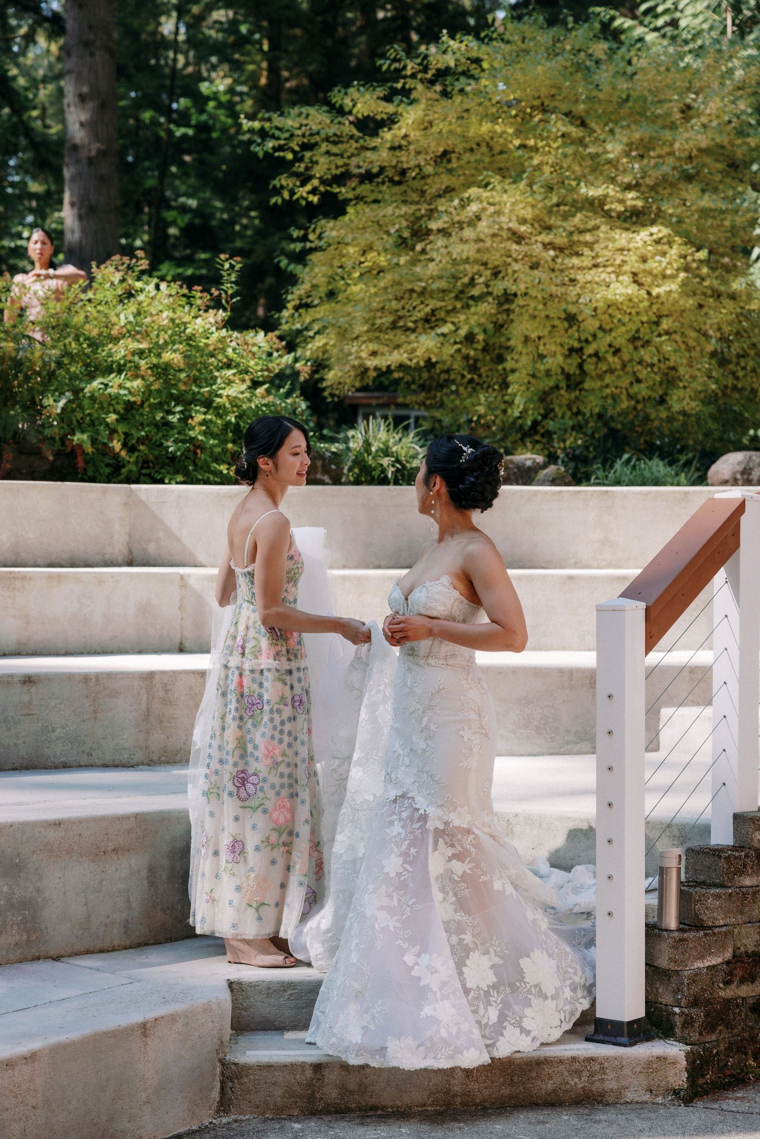 Bridesmaid in floral dress helping bride in lace wedding gown on stone steps at outdoor venue.