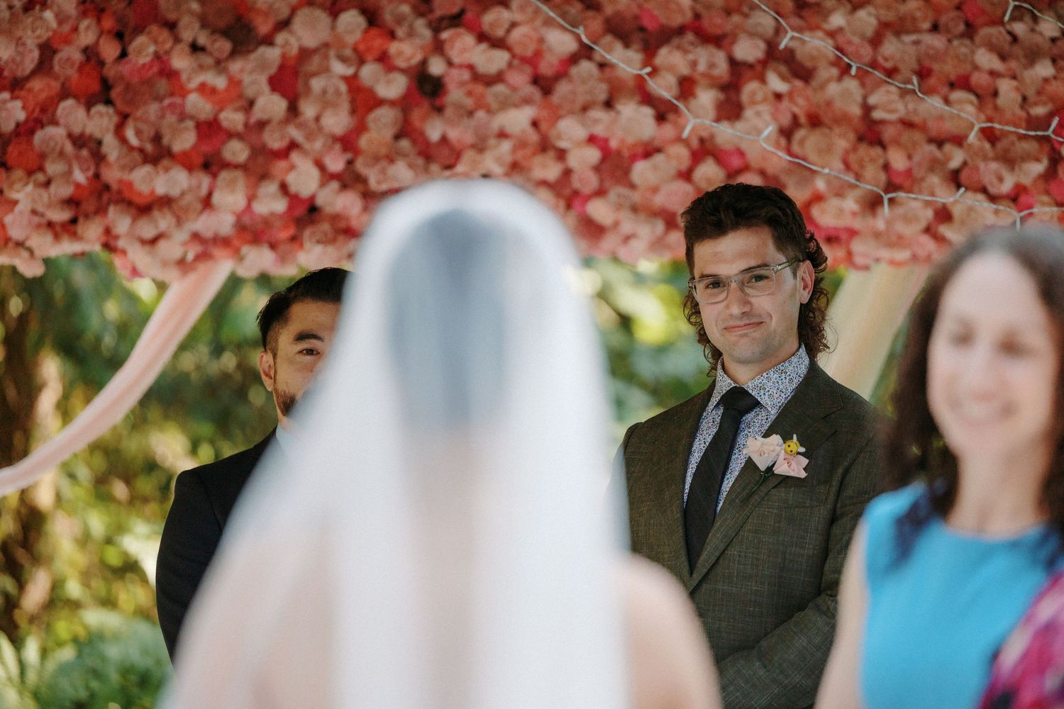 Groom in grey suit watches bride approach at outdoor ceremony beneath pink floral wedding arch.