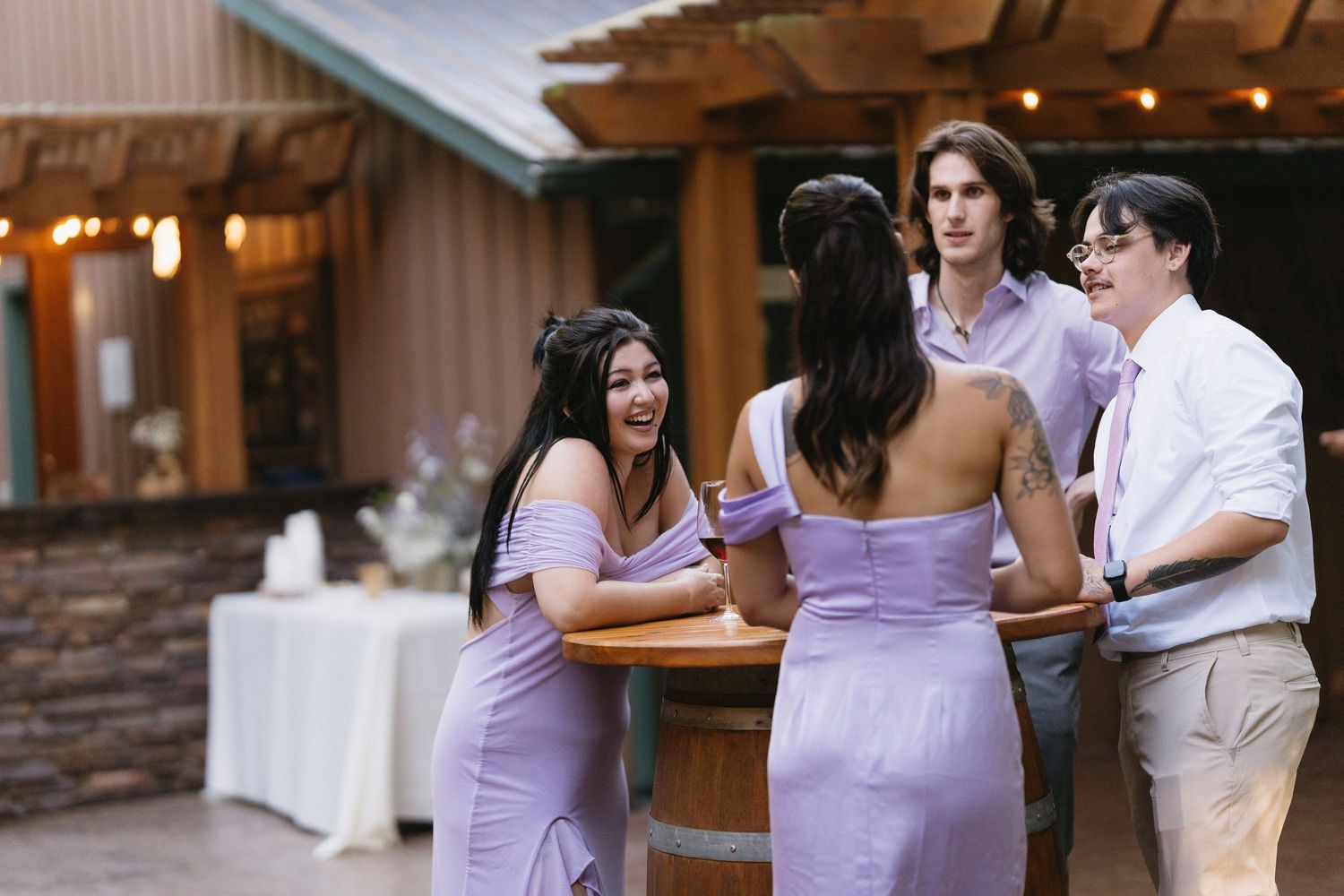 Bridesmaids in lavender dresses socialize with guests around a barrel table at outdoor wedding.