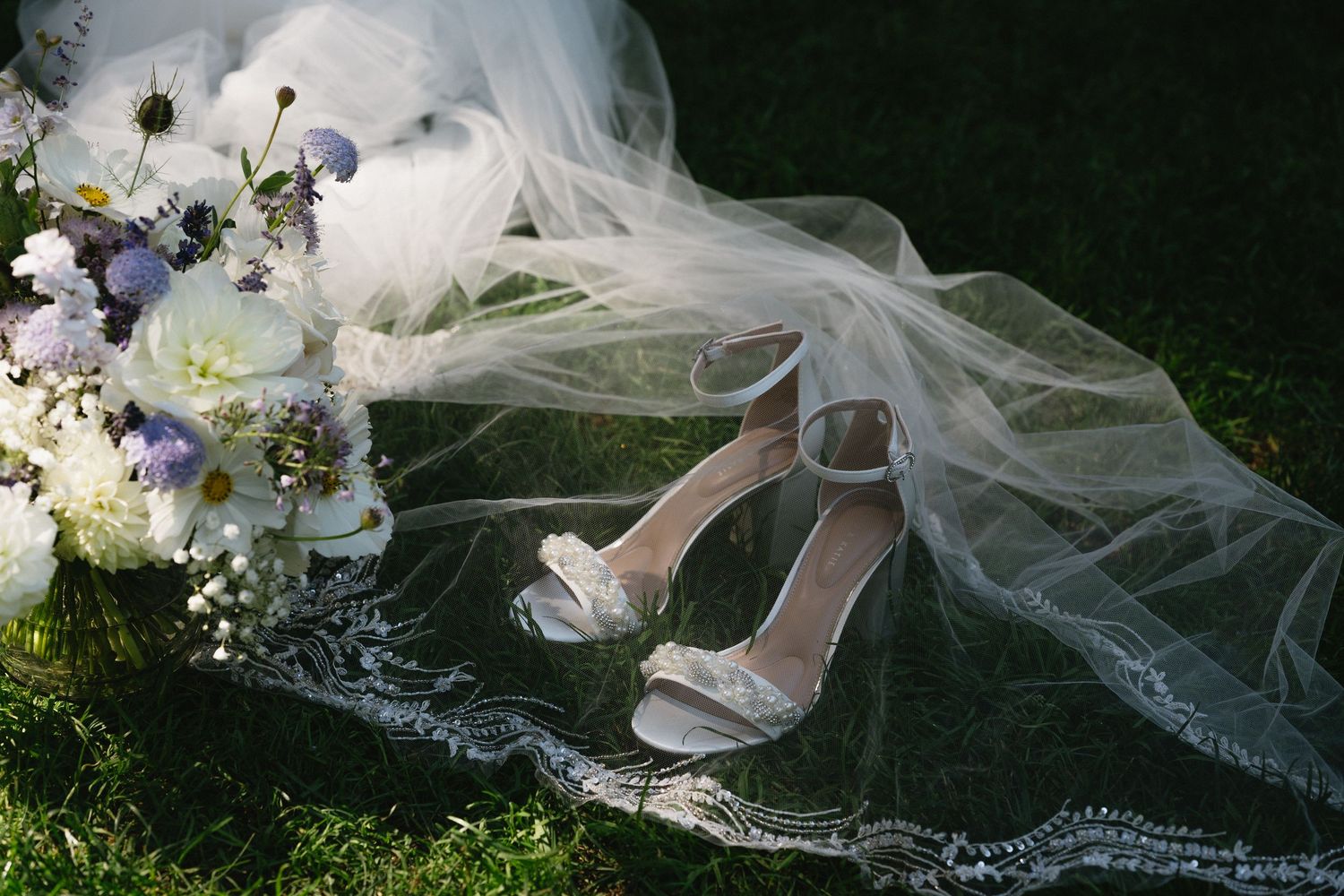 Bridal heels and bouquet arranged on grass beneath a lace-trimmed wedding veil.