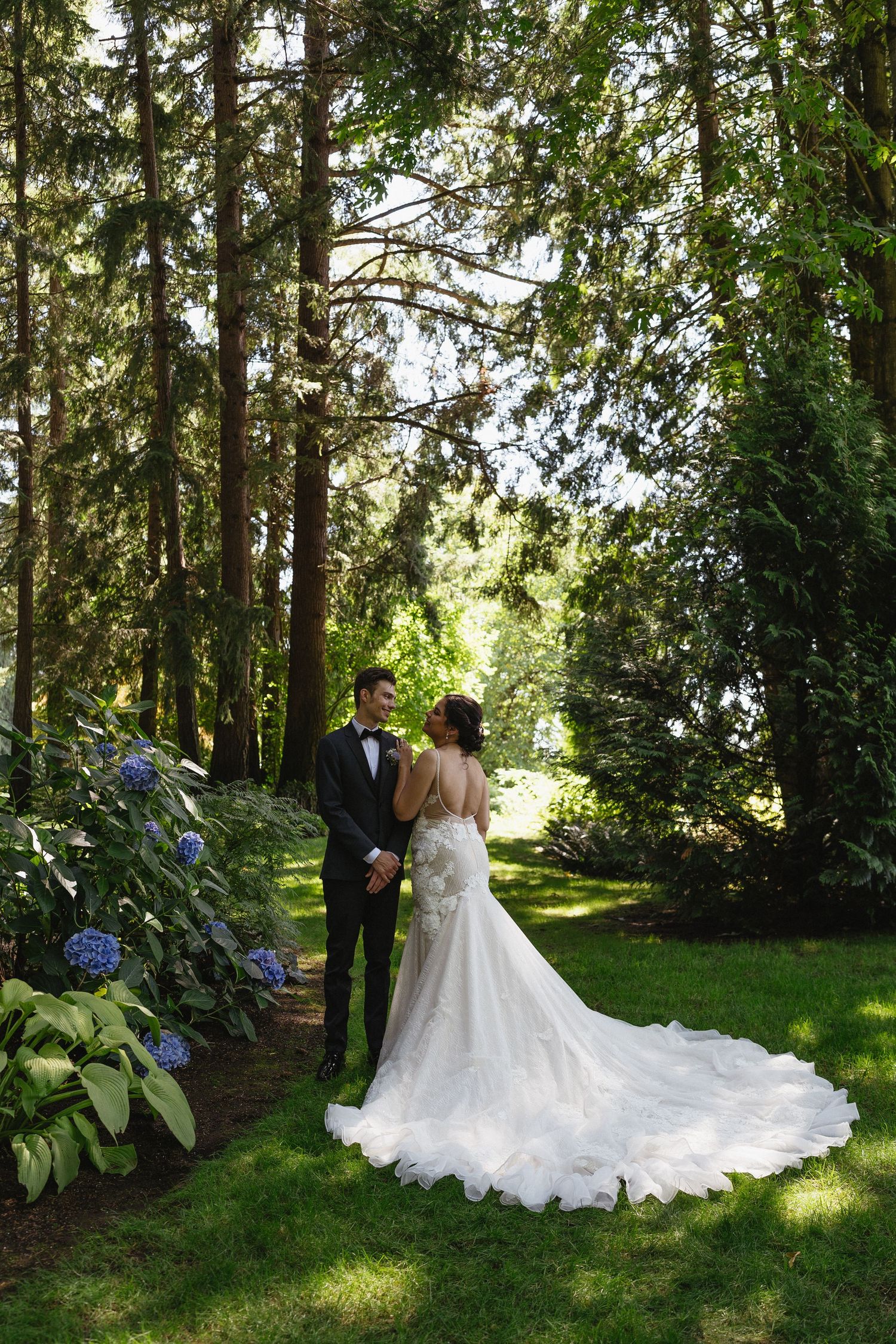Bride and groom walk hand in hand through a lush garden path lined with hydrangeas and tall evergreen trees.