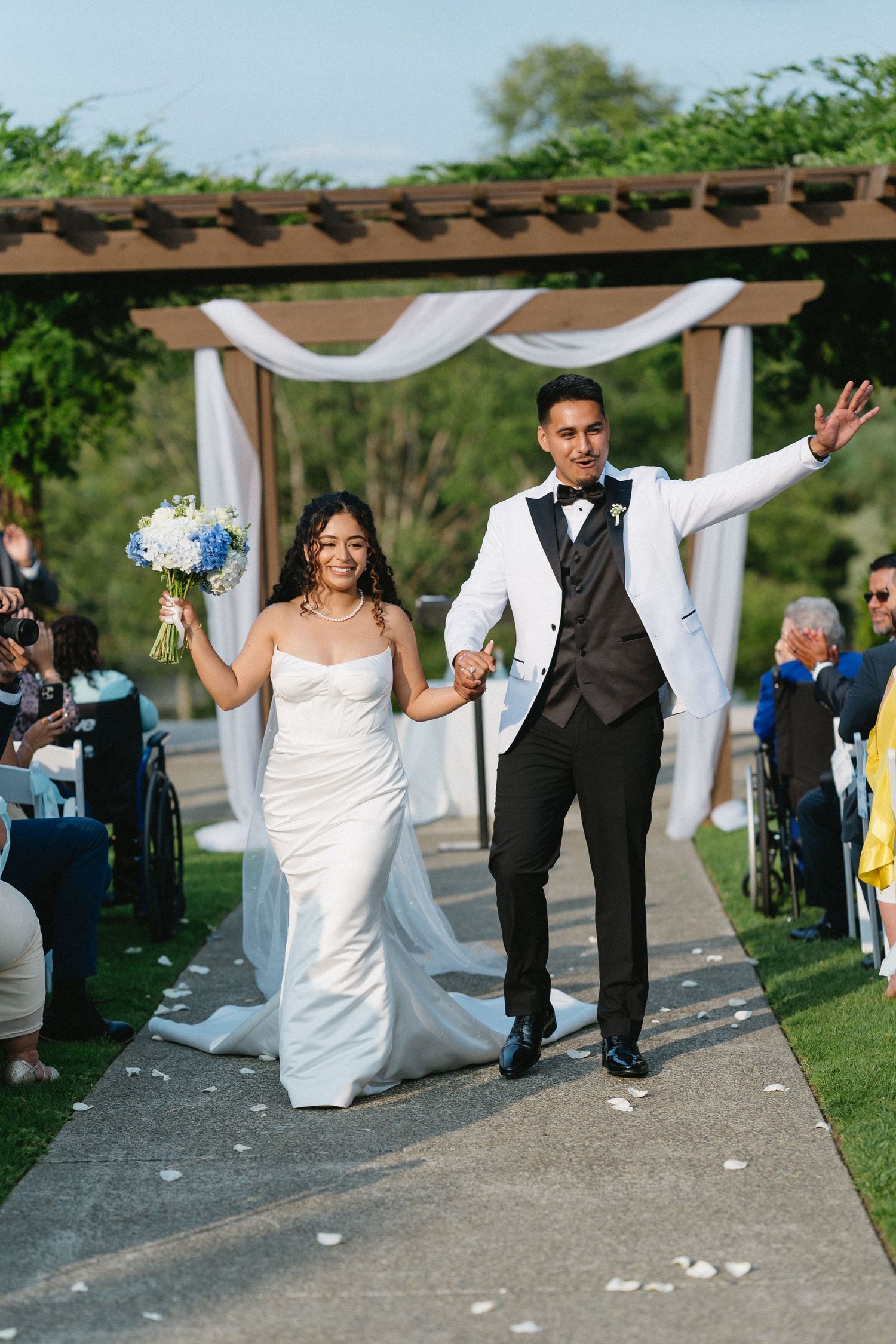 Newlyweds walk down aisle holding bouquet, waving to guests after outdoor ceremony.