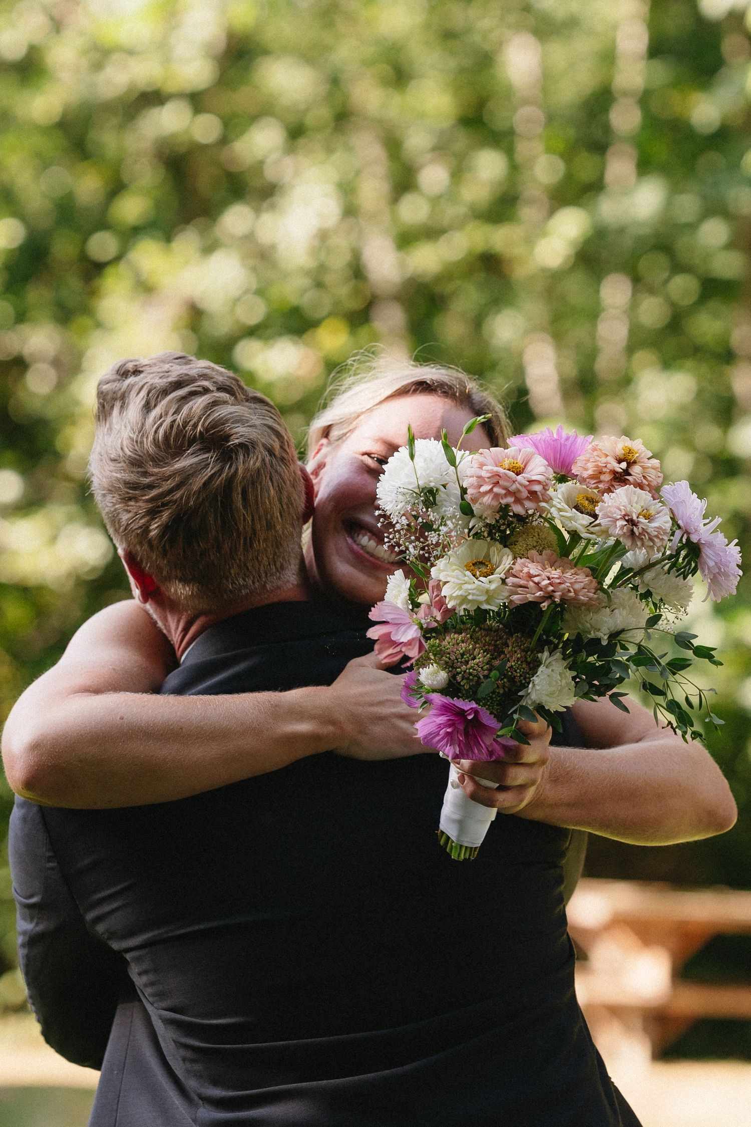 Joyful woman holding colorful bridal bouquet sharing warm embrace with groom at outdoor wedding.