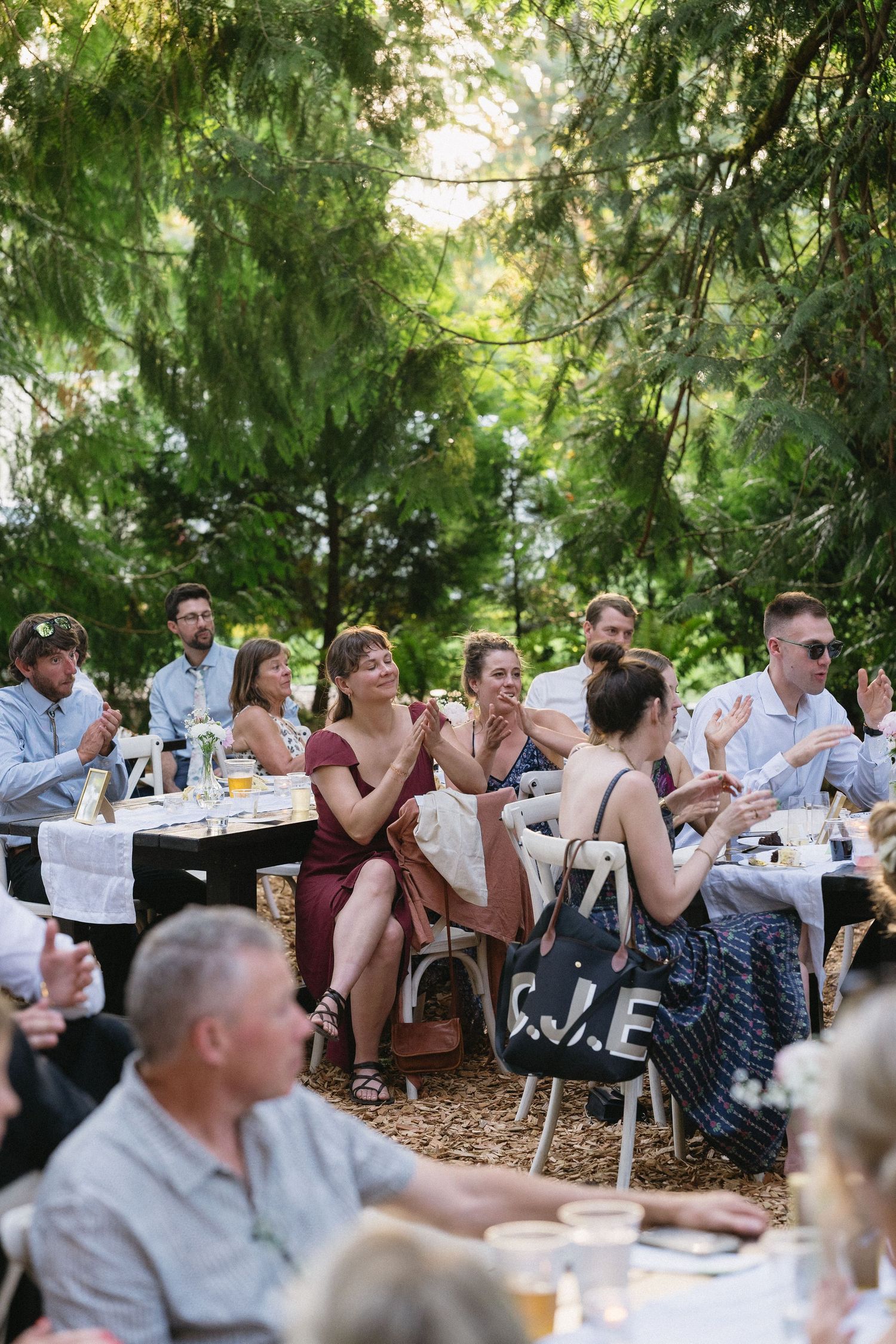 Wedding guests clapping and smiling at an outdoor garden reception surrounded by lush green trees.