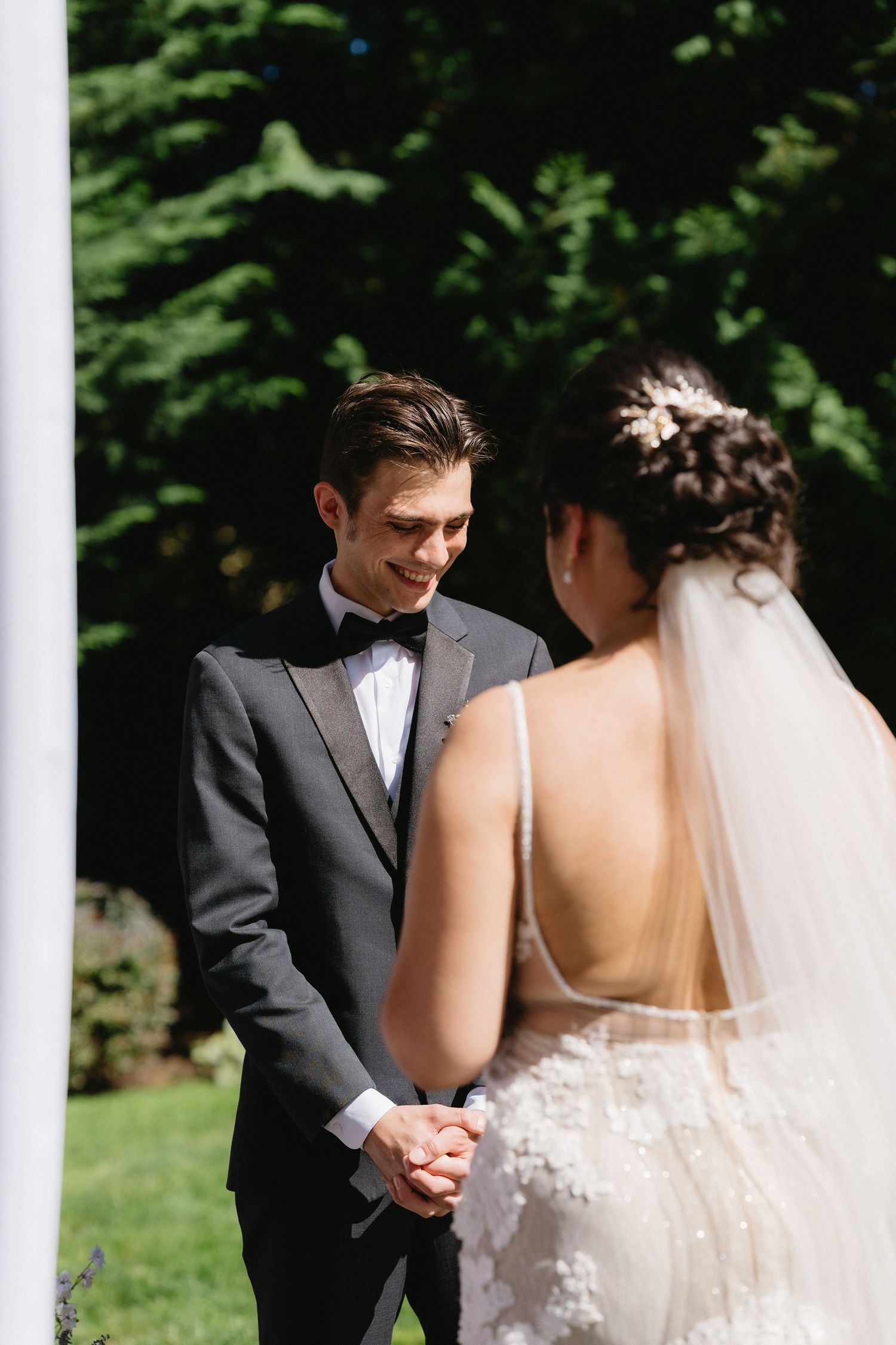Smiling groom in gray tuxedo holds bride's hands during outdoor wedding ceremony among tall green trees.
