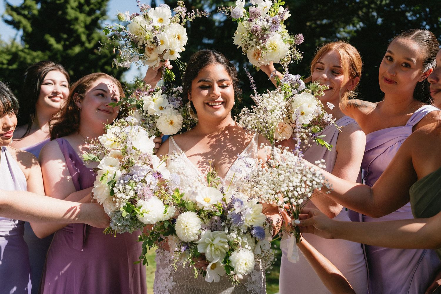 Bride surrounded by bridesmaids in purple dresses holding white floral bouquets outdoors.