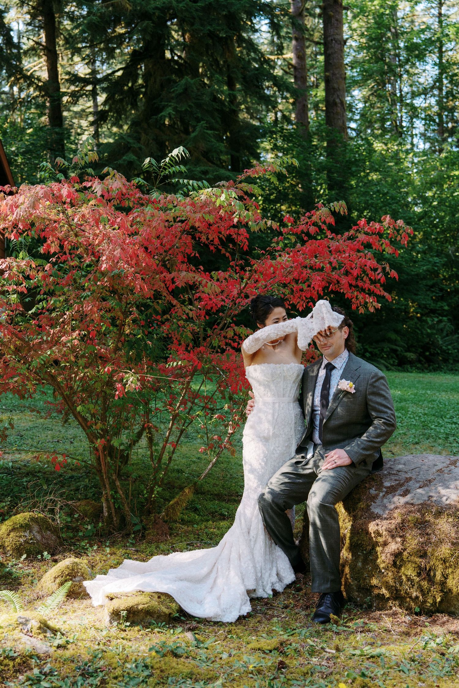 Bride and groom pose by vibrant red Japanese maple tree on mossy rocks in a lush forest wedding setting.