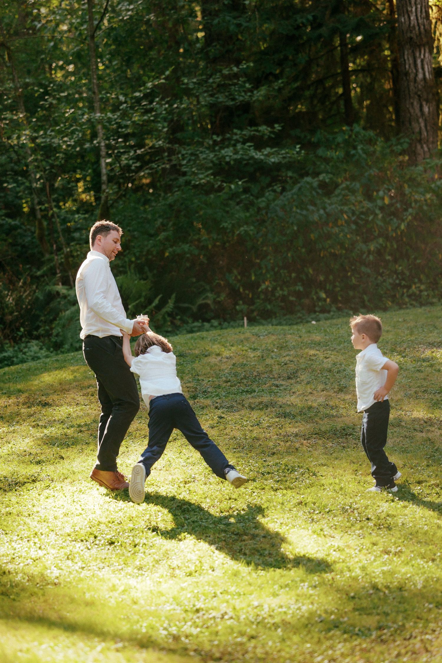 Man in white shirt playfully swings a child while another boy watches on sunlit green lawn at wedding.