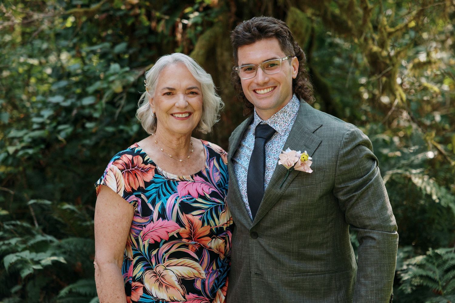 Groom in gray suit poses with older woman in floral dress at outdoor wedding.