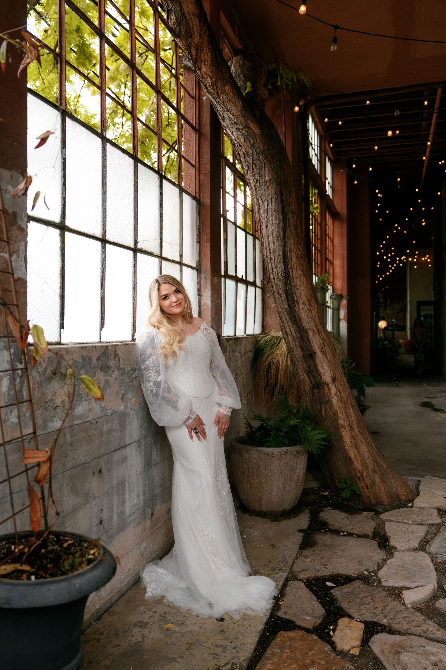 Elegant bride in long-sleeve lace wedding gown posing beside a tree and large windows with fairy lights.