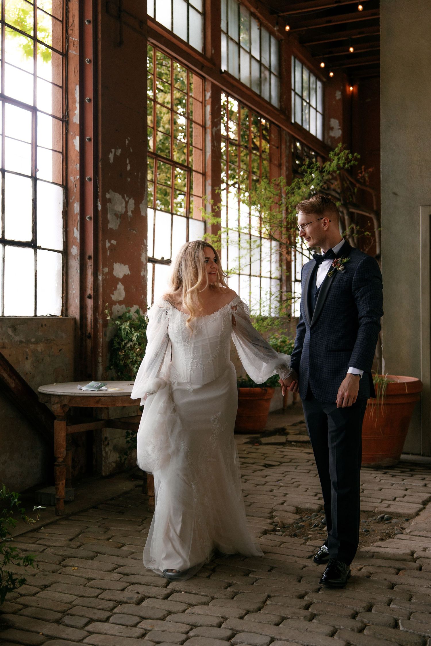 Bride in off-shoulder lace gown and groom in navy tuxedo walk hand in hand through sunlit industrial wedding venue.
